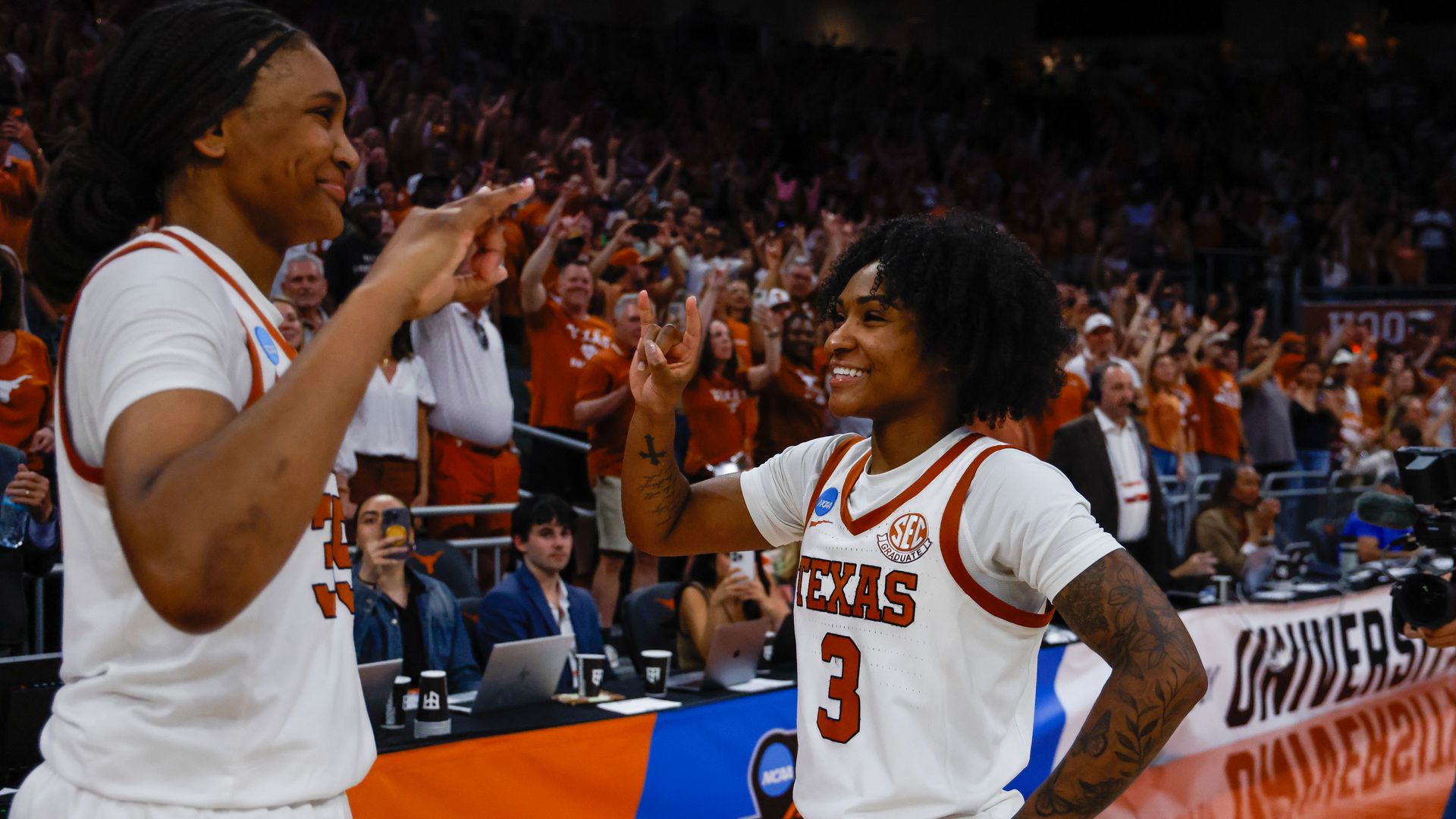 Two Texas women's basketball players in white jerseys high-five on the court as a crowded arena cheers, fans in orange visible in the stands.