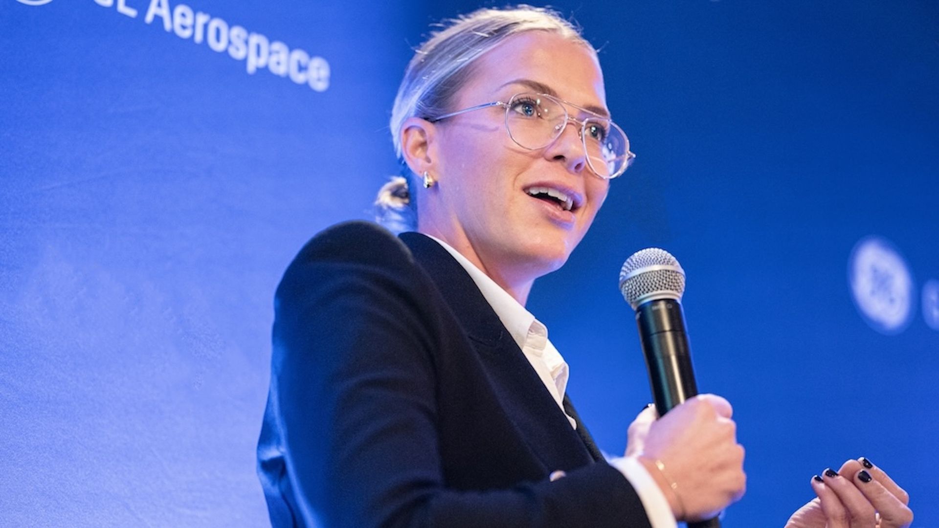 A woman speaking into a microphone in front of a blue background.