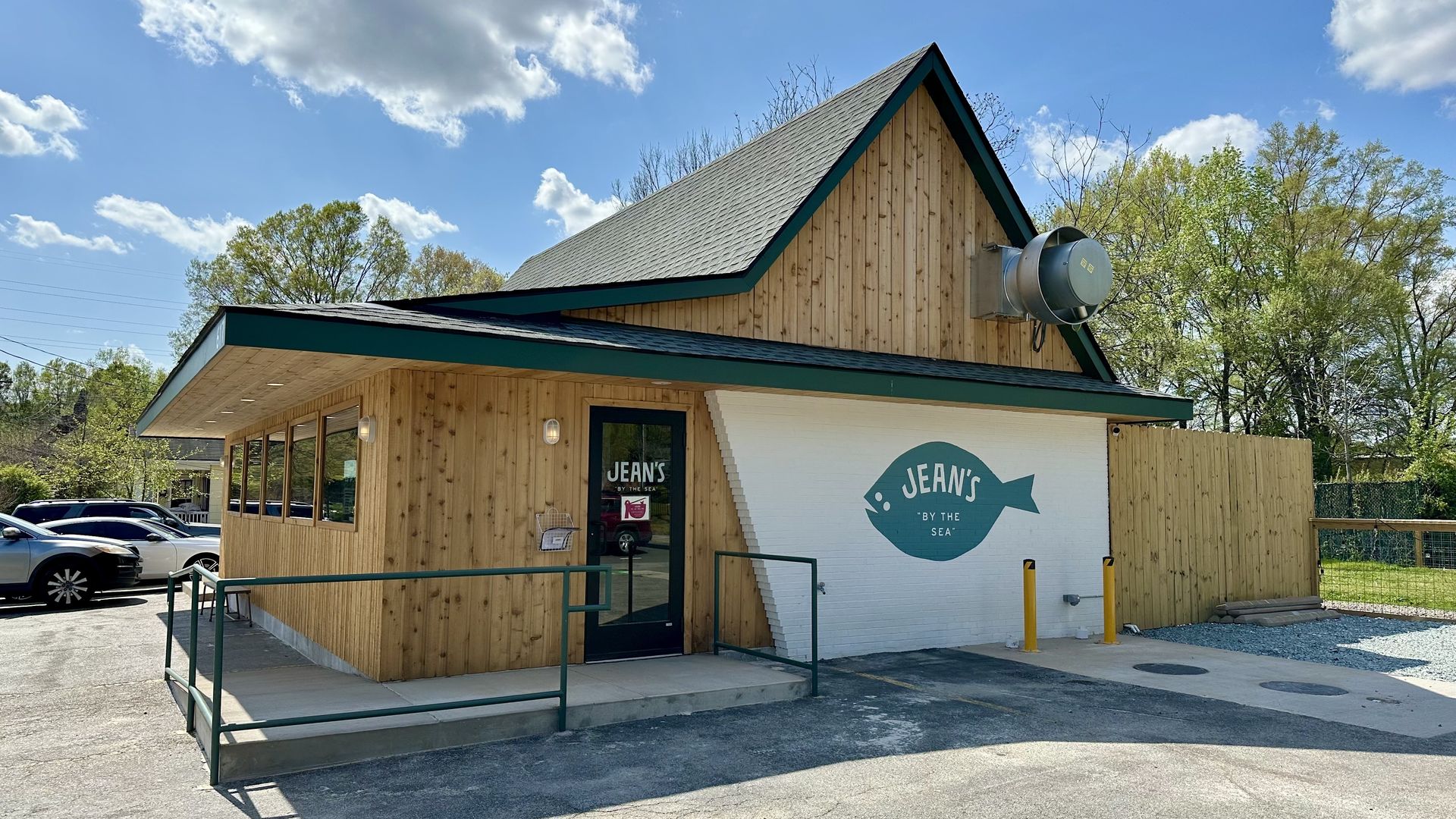 Wooden A-frame cafe with teal trim. White wall shows a teal fish logo and "JEAN'S BY THE SEA". Parking lot with cars, green railing, yellow bollards, blue sky and trees behind.