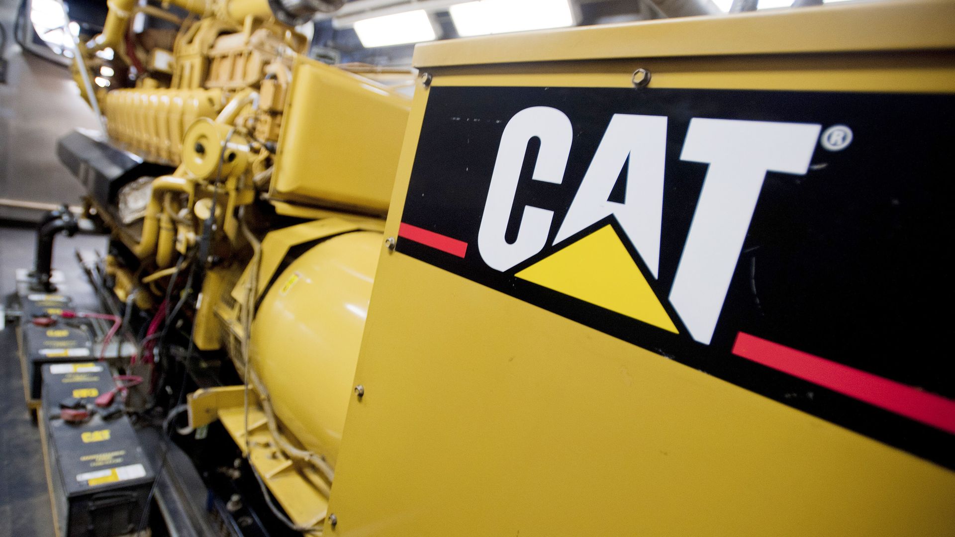 Close-up of a yellow Caterpillar machine with a large black panel displaying the white CAT logo; visible engine components, hoses, and a battery pack in an industrial workspace.
