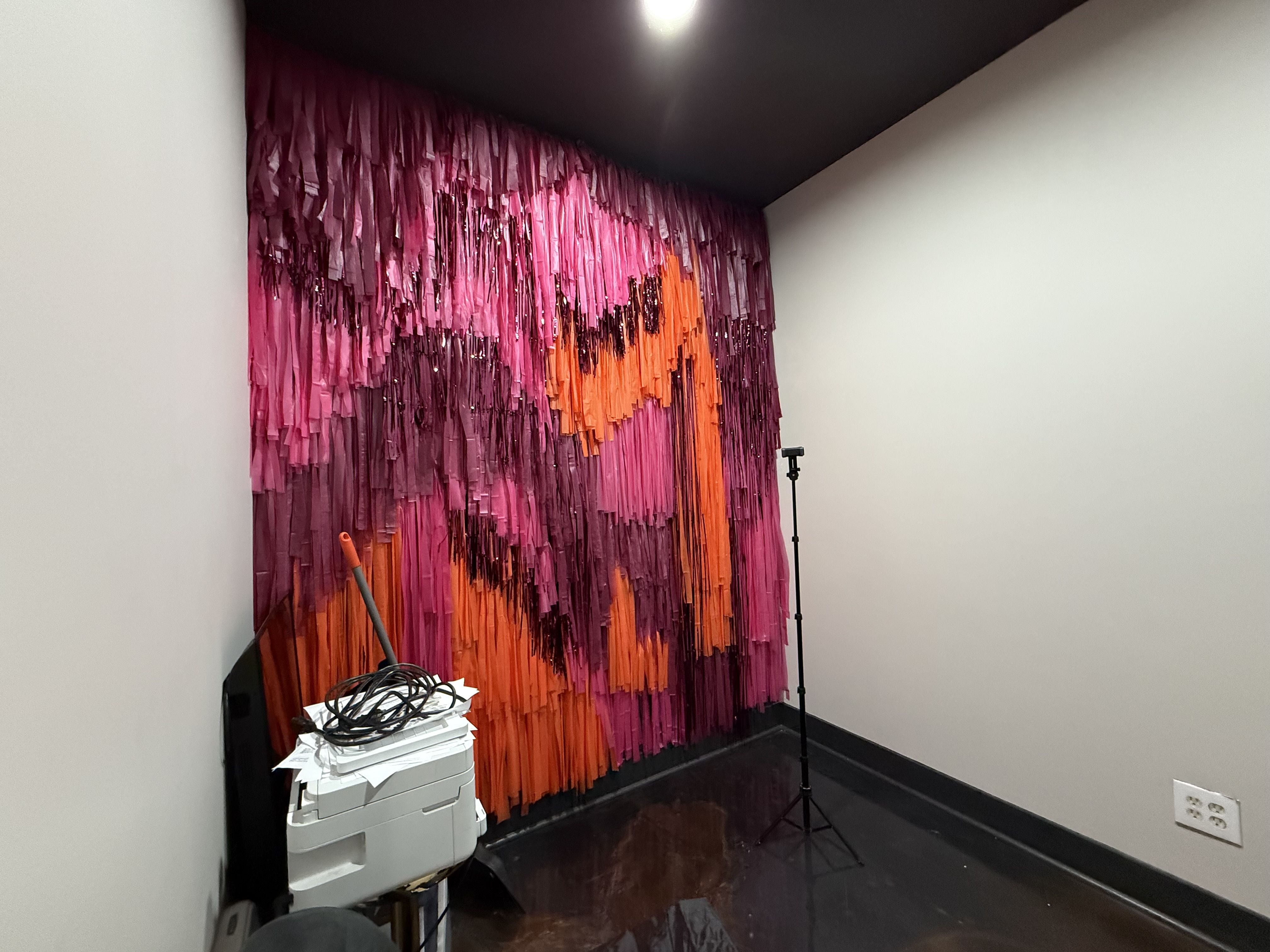 Corner room with a wall covered in pink and orange fringe strips forming an abstract gradient. A tall lighting stand and tangled cables sit near a white printer on the dark glossy floor.