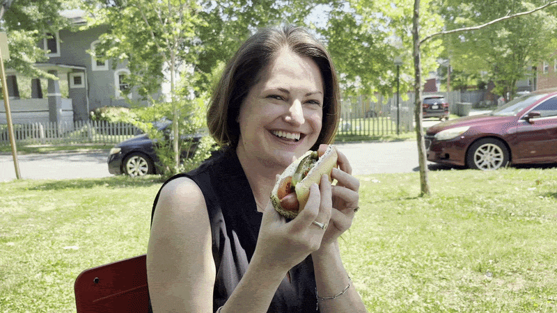 Woman wearing a black top biting into a hot dog in a park with green grass and houses in the background.