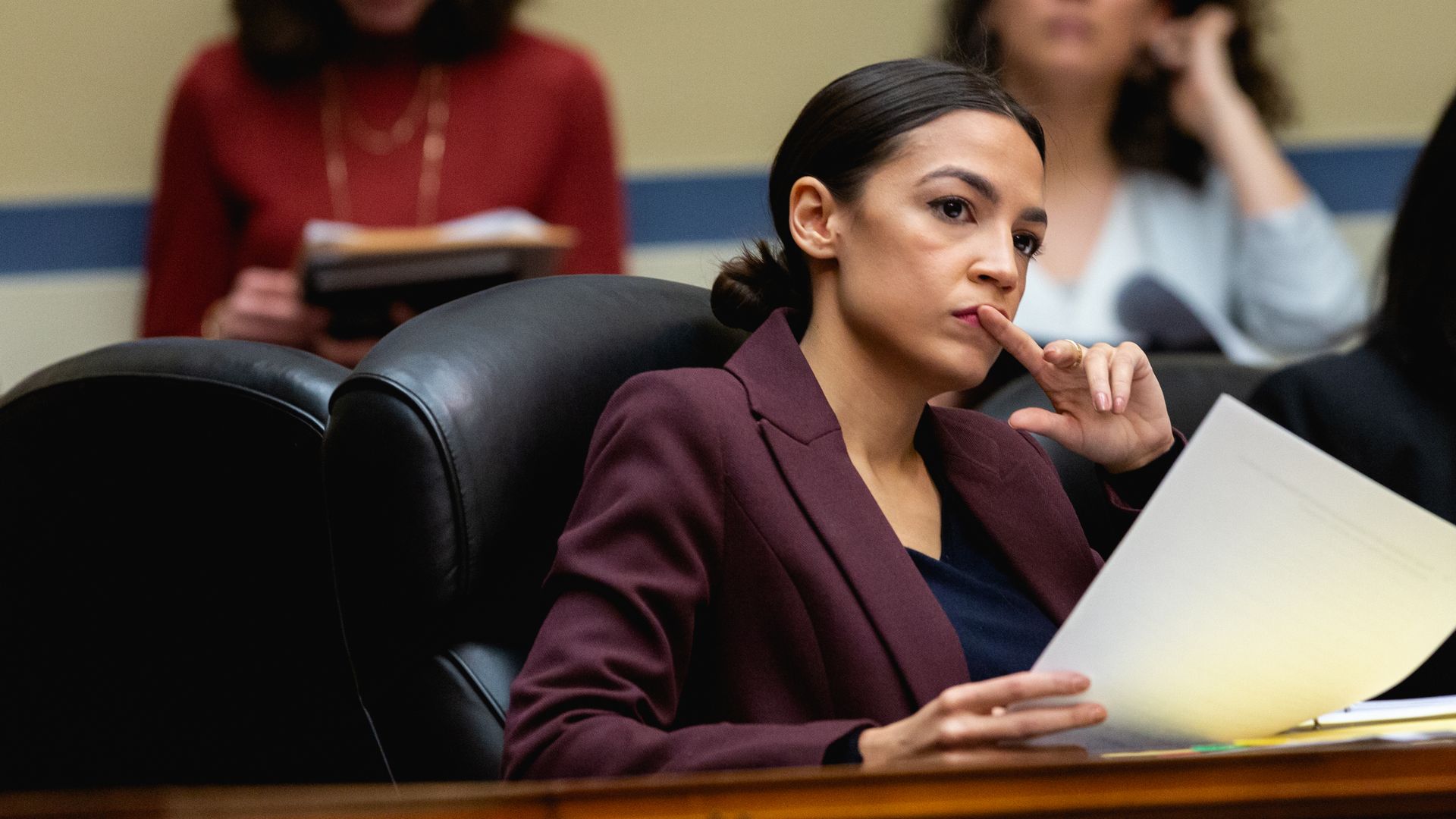 AOC looking intensely forward with her hand touching her mouth. 