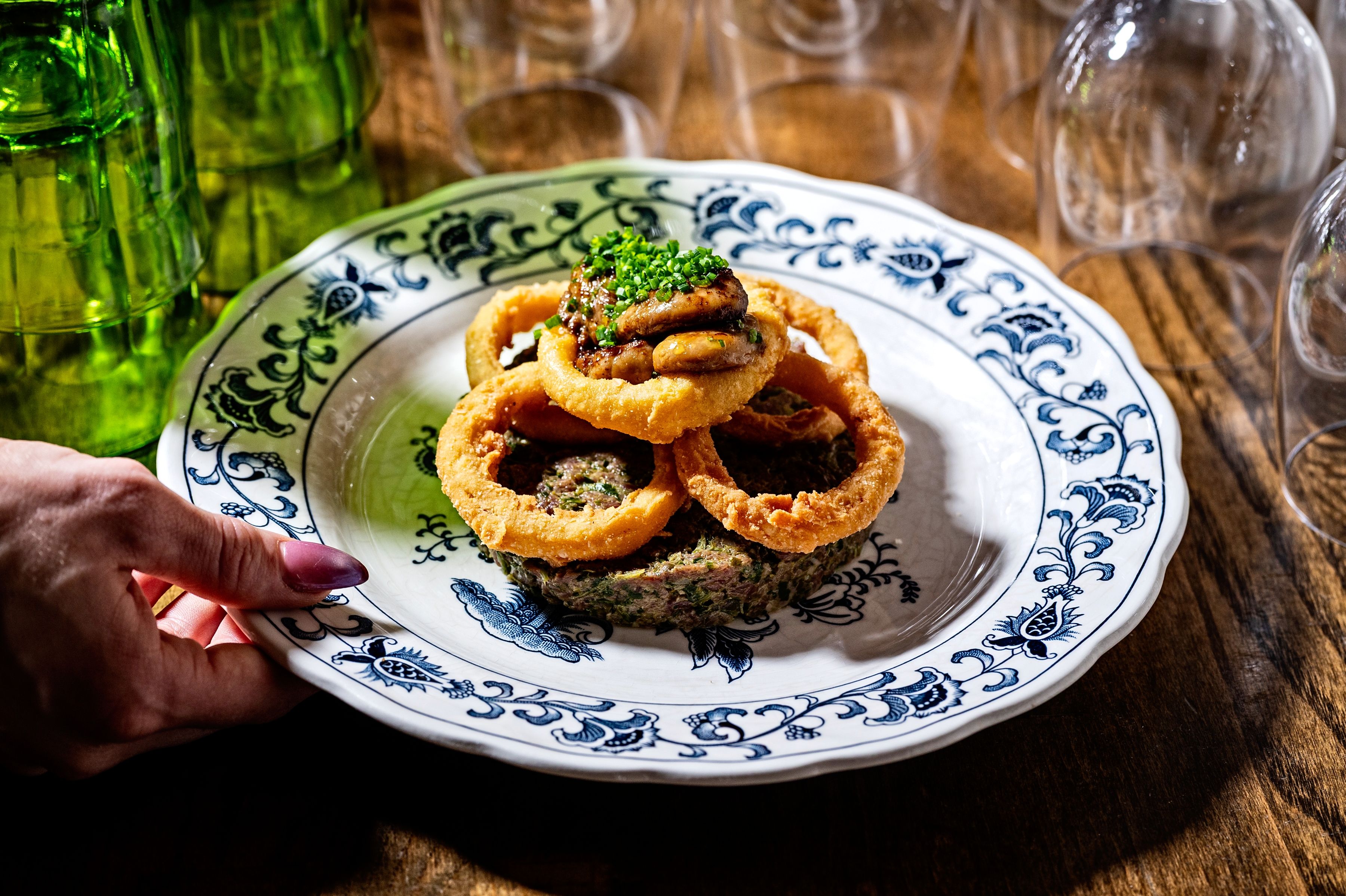 Blue and white patterned plate with a stack of onion rings around a mound of lamb tartare, topped with mushrooms and chopped chives. A hand holds the plate; green bottles and glassware blur in the background.