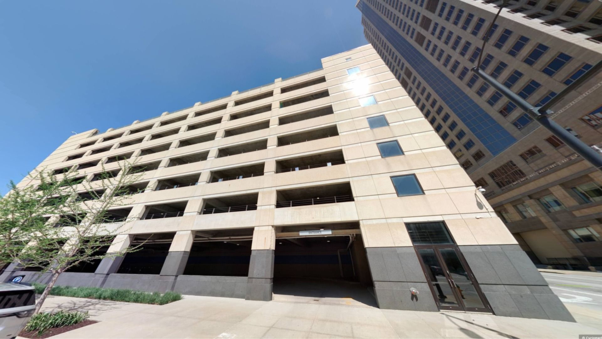 Looking up at a multi-level concrete parking garage with open sides under a bright blue sky beside tall office buildings on a sunny day.