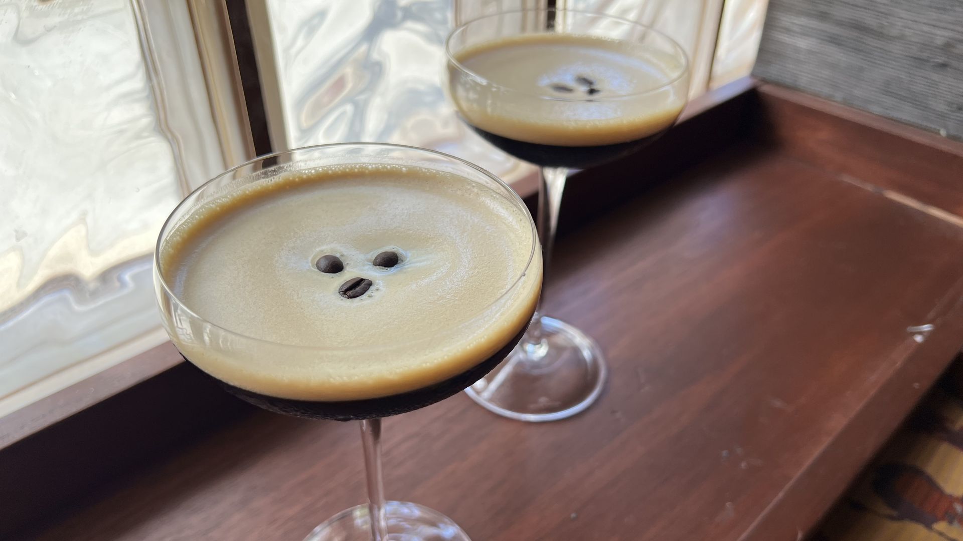 Two coupe glasses sit on a wooden bar by a window, each filled with an espresso martini decorated with three small coffee beans in the middle at Jue Let.