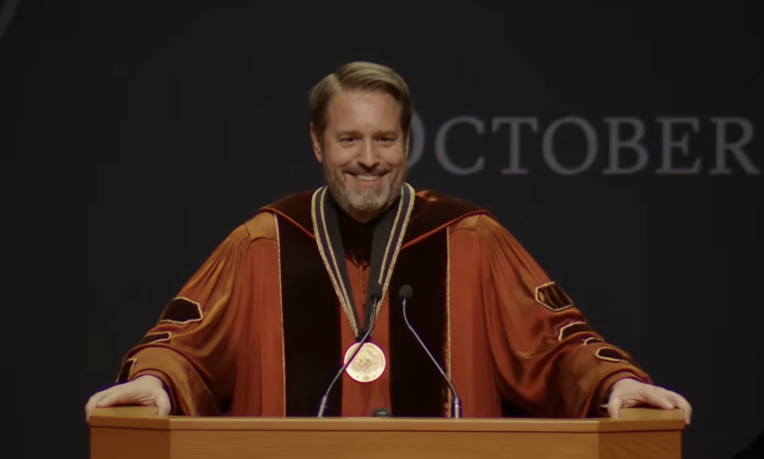 Smiling man with light brown hair and beard in orange and black academic gown stands behind a podium with two microphones, with part of the word "OCTOBER" visible on black background.