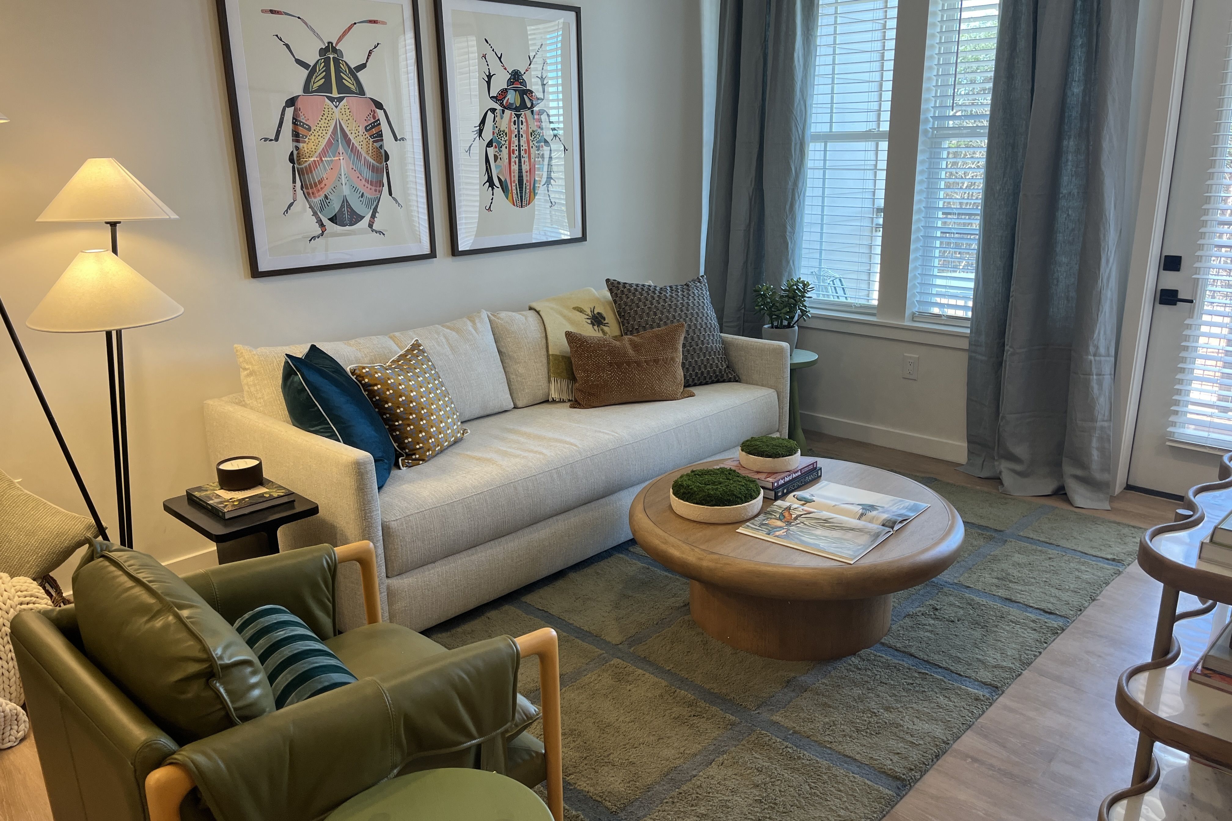 Living room with a light gray sofa and colorful pillows; two framed beetle prints above. Round wooden coffee table with moss bowls and magazines. Large window with blue curtains.