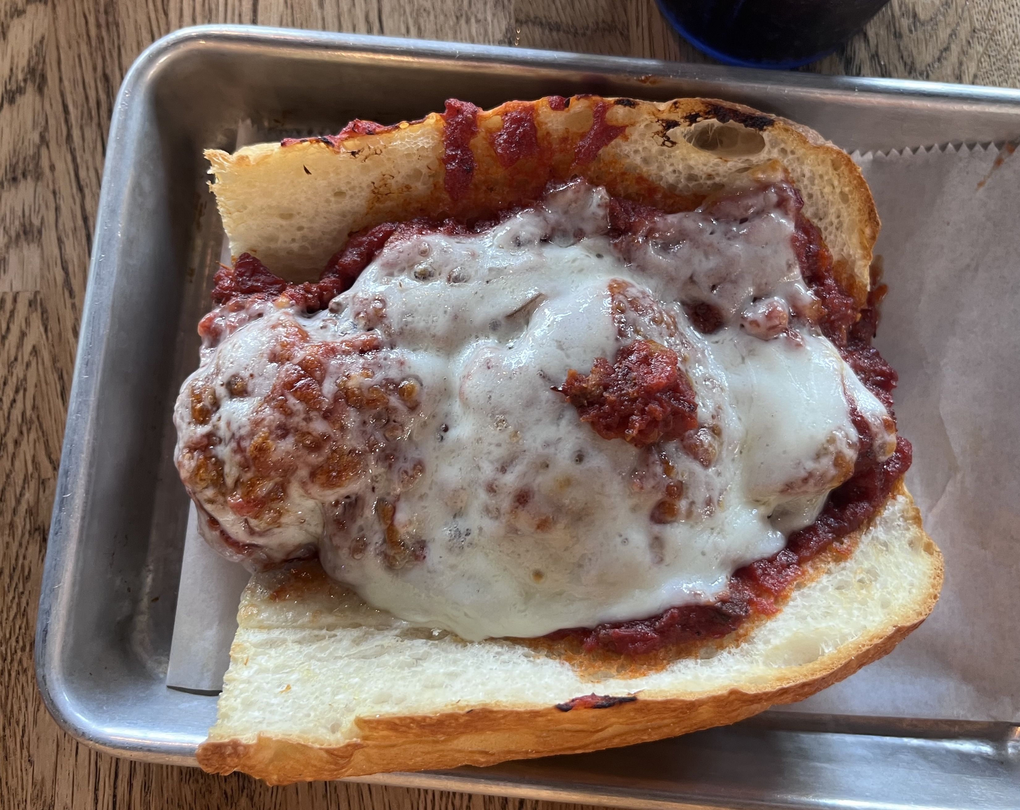 A thick slice of bread topped with meatballs and melted cheese in a metal baking pan; crusty edges, red sauce visible, on a wooden table.