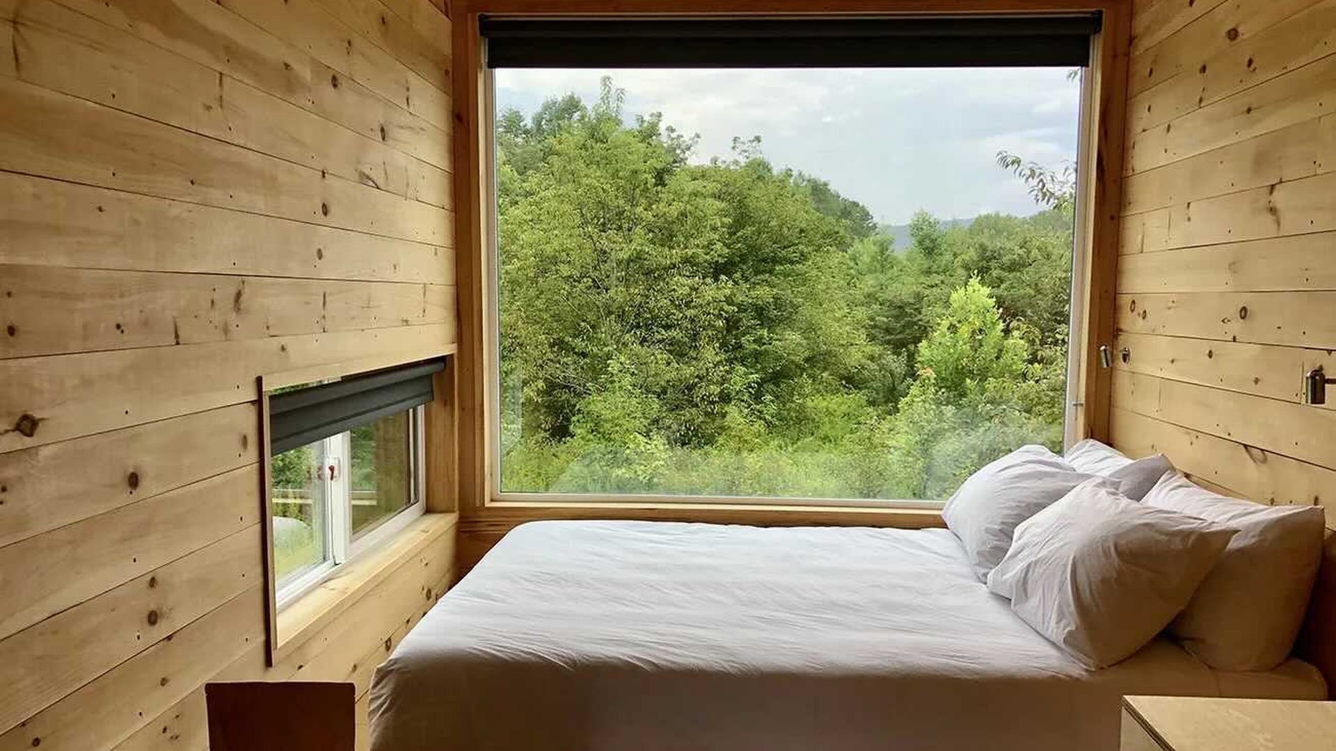 Queen sized bed in front of large window overlooking woods in summer
