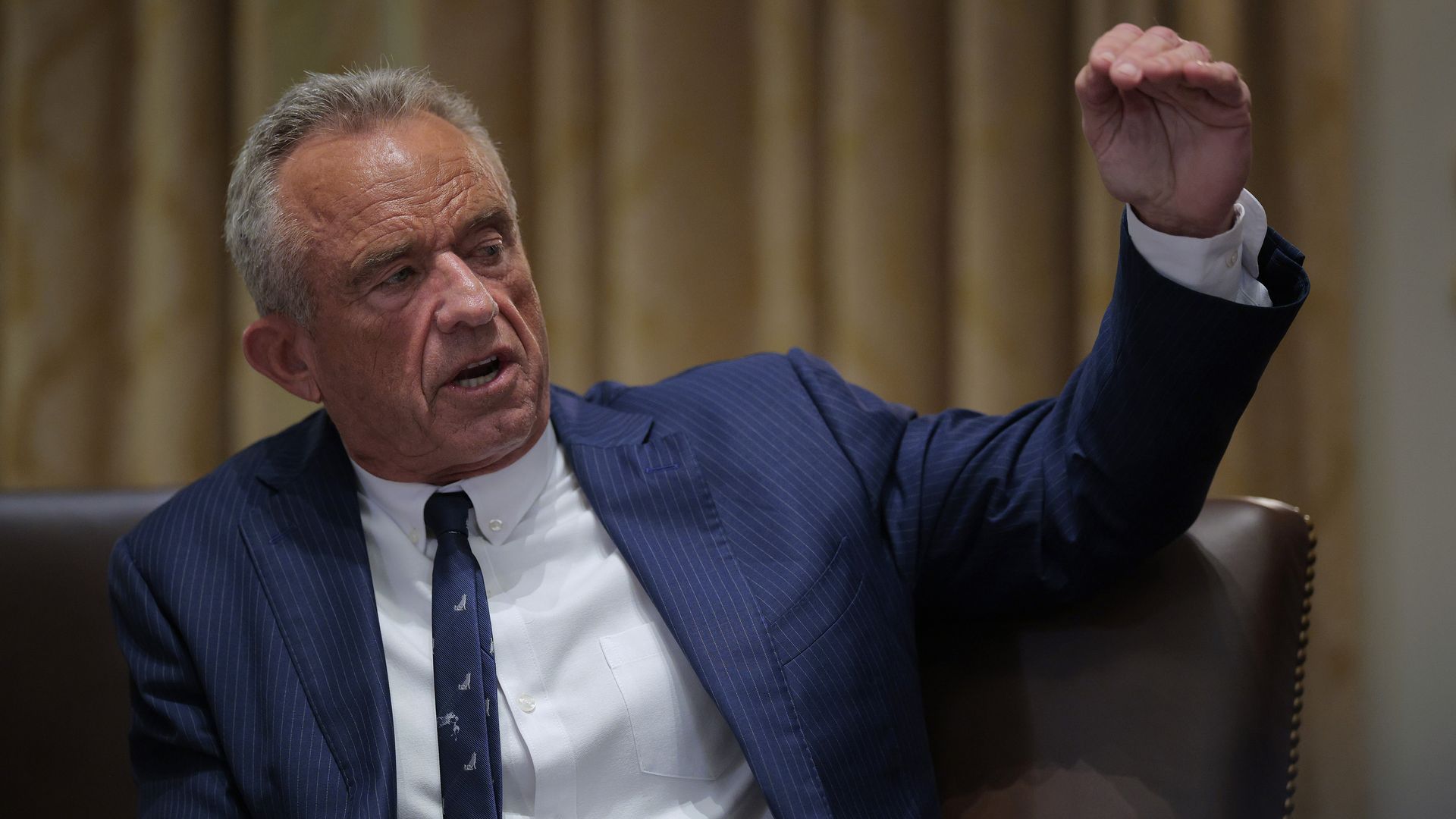 RFK wears a blue suit, white shirt and blue tie. He's speaking with one hand outstretched in front of him.