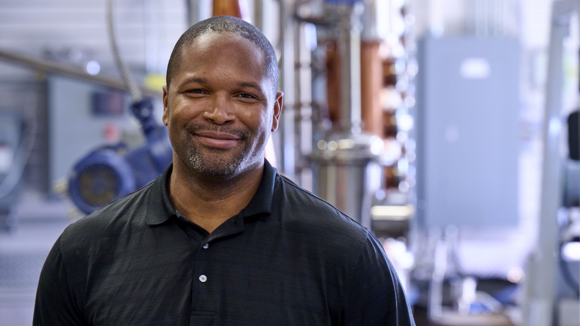 Chris Montana poses in front of distilling equipment. He's wearing a black polo.