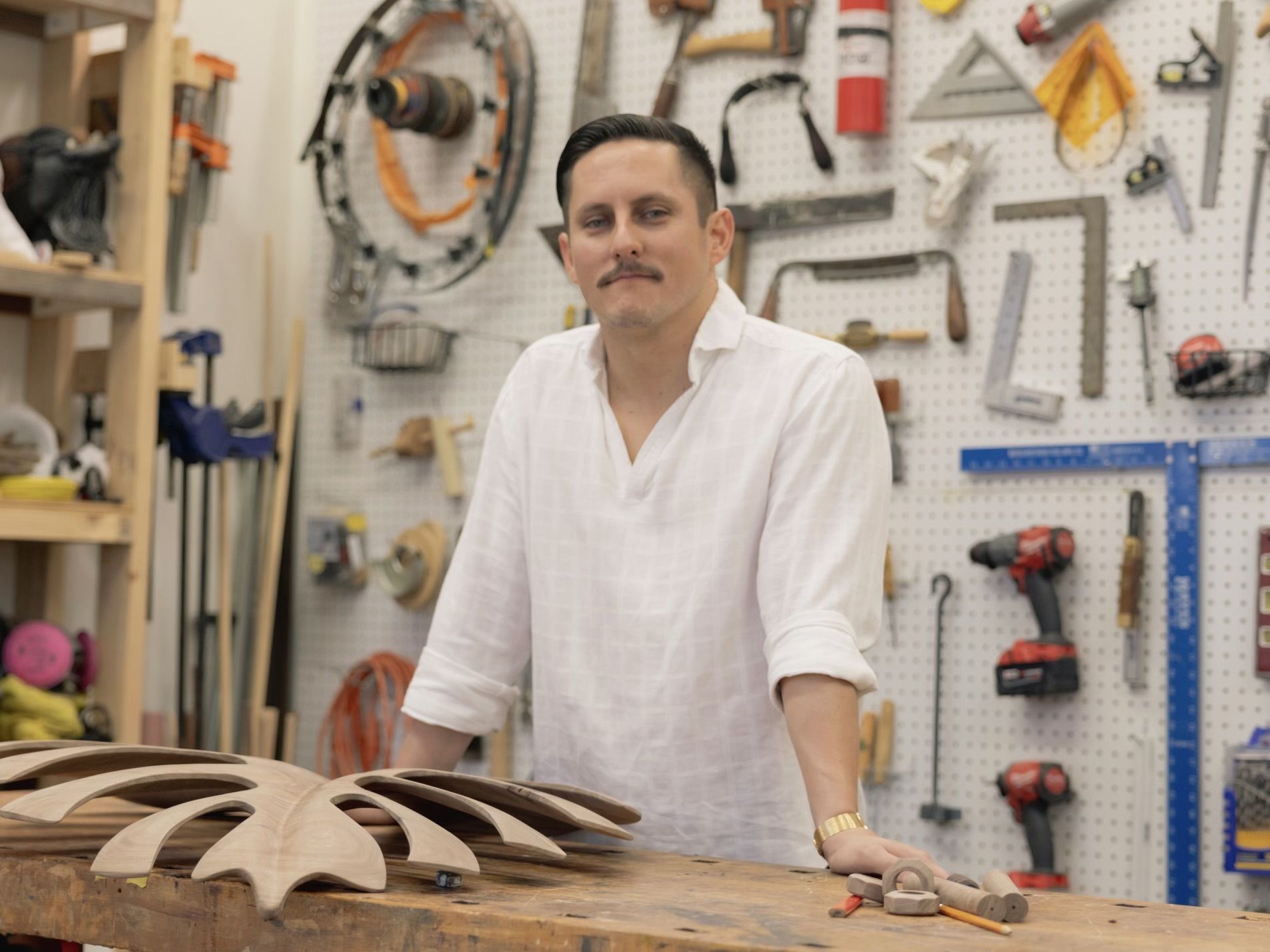 Man in a white shirt with rolled sleeves stands at a wooden workbench with a carved wooden piece resembling a flower, tools and equipment hang on a pegboard behind him.