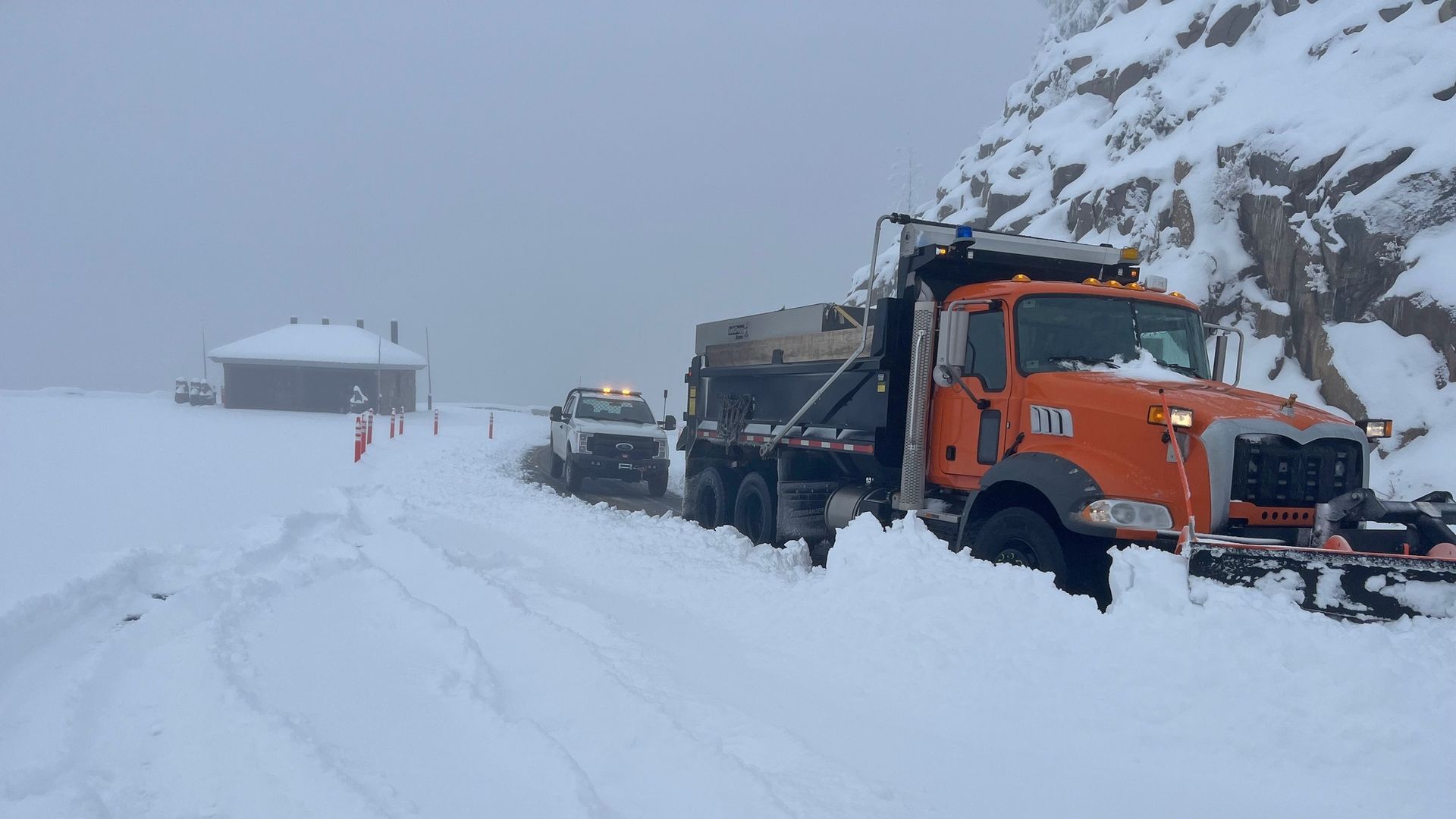 Snowy mountain road with an orange heavy-duty snowplow truck in the foreground and a white pickup behind it. Snowbound hut, rocky cliff to the right, and orange traffic cones along the road.