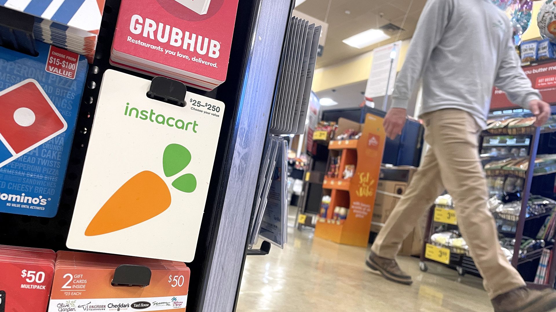 Gift cards for Domino's, Grubhub, Instacart, and Dinner and a Movie displayed in a store aisle with a person walking past in beige pants and brown shoes.
