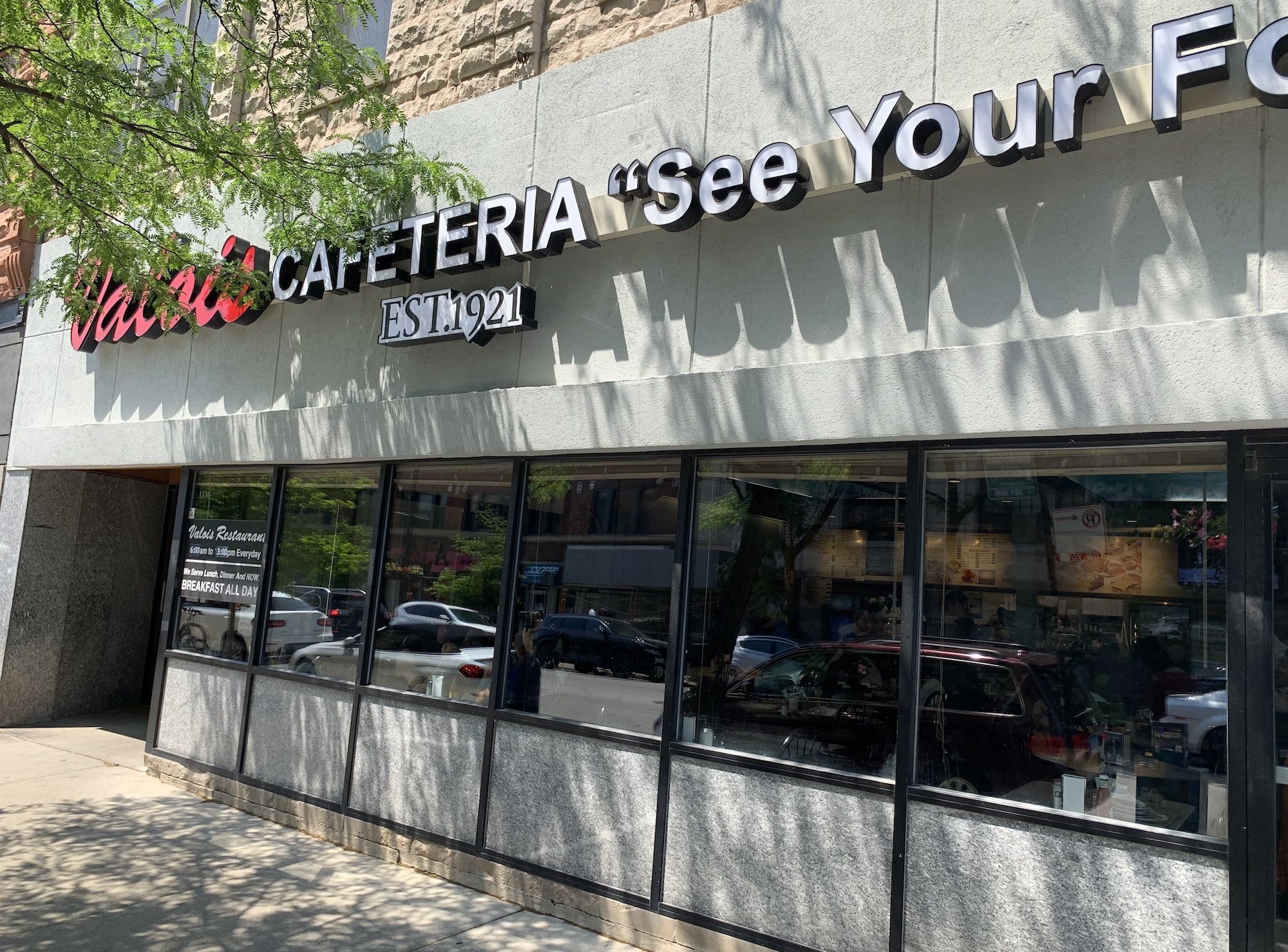 Exterior storefront with large 3D sign CAFETERIA 'See Your F' and EST.1921 on a gray wall; glass windows, reflections of cars, and tree shadows.