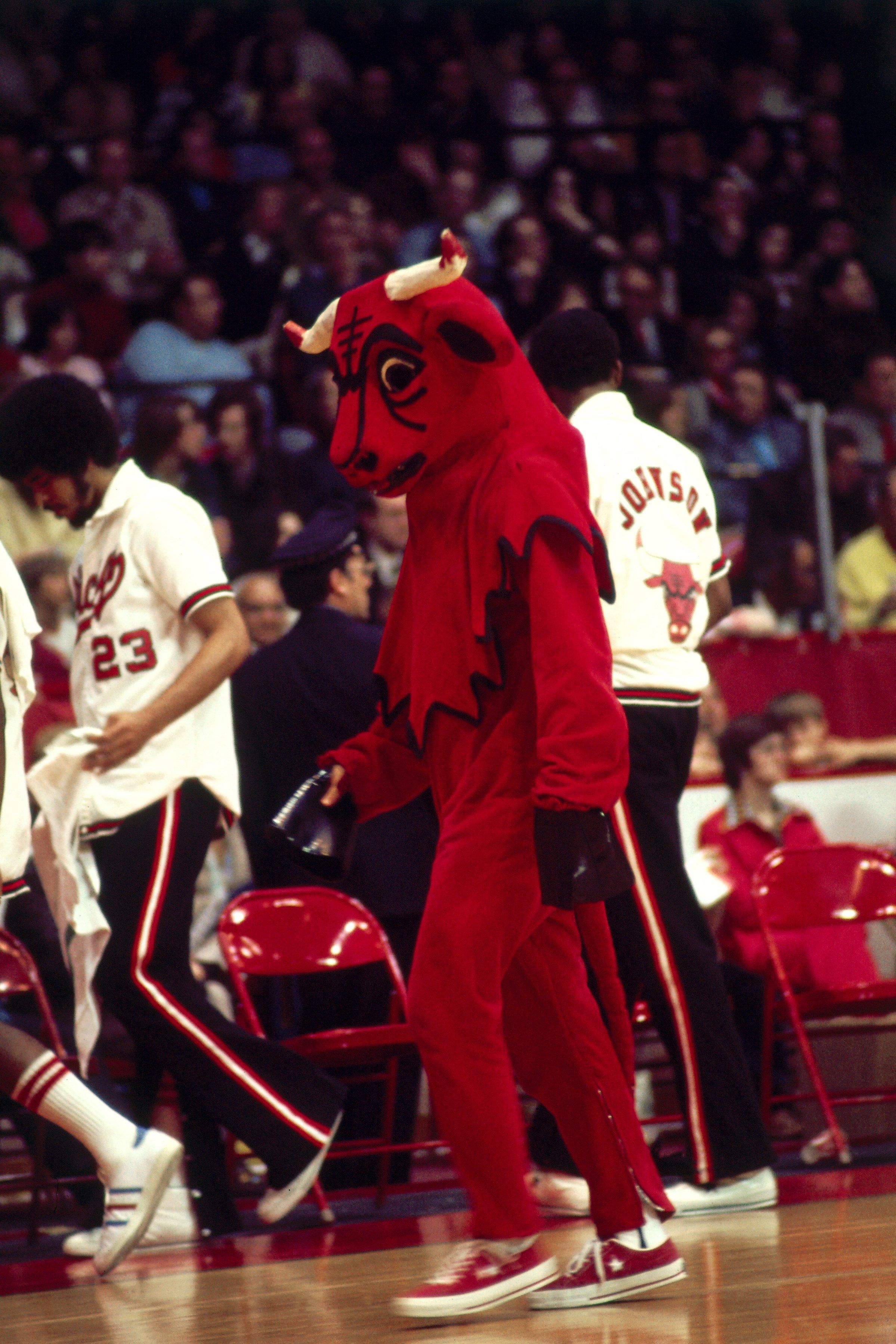 A red bull mascot with horns and black markings walks on a basketball court, surrounded by Chicago Bulls players in white uniforms and a cheering crowd in the stands.