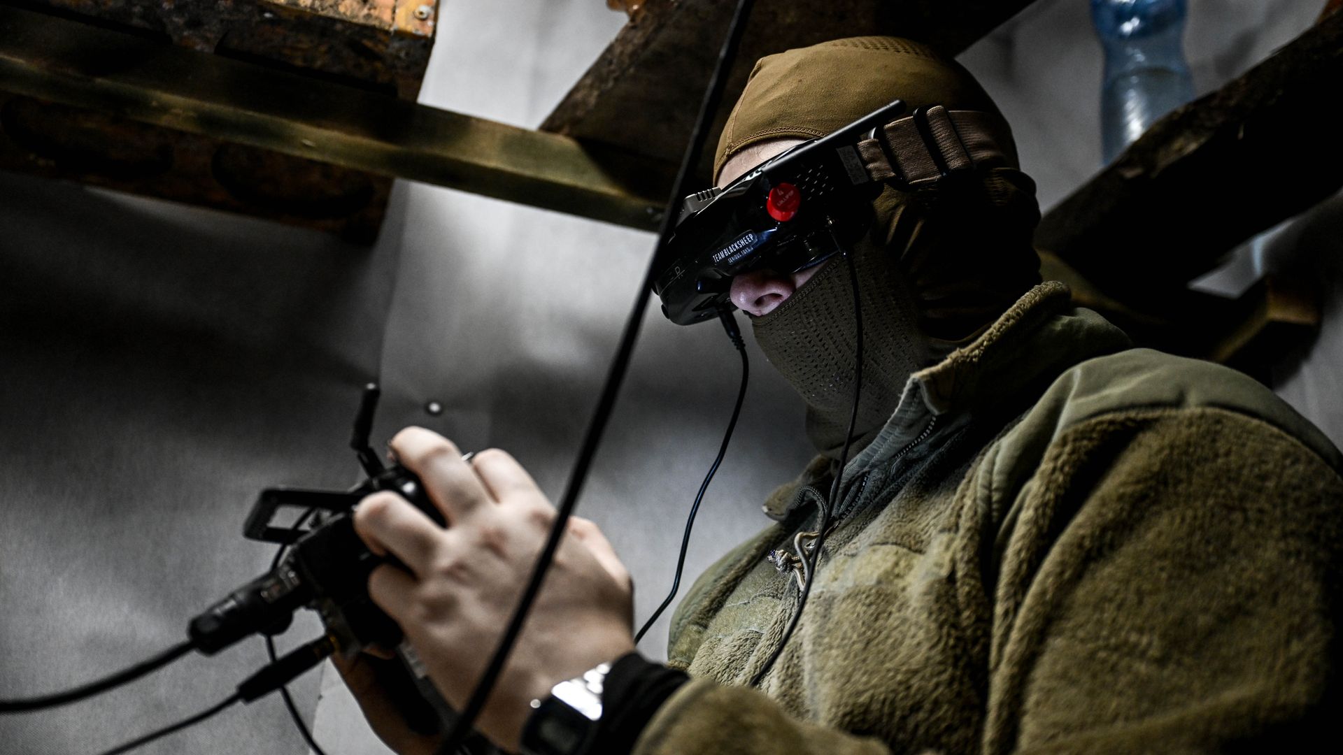 A man sits in a dugout, operating an FPV drone. He holds a control and wears a futuristic headset. Shelves and random items surround him.