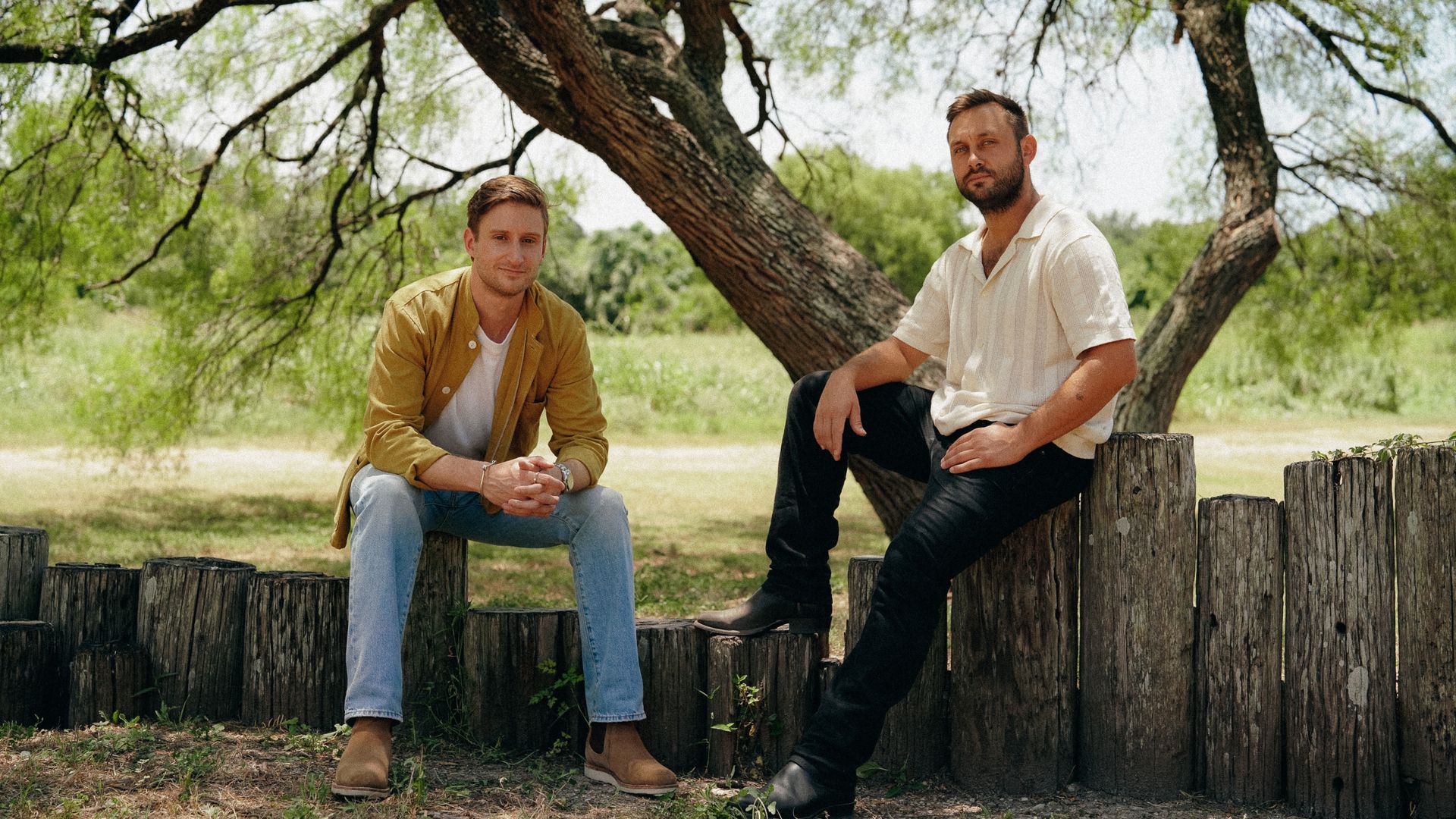 Two men sit outdoors on wooden logs under a tree with green leaves. One wears a yellow jacket, white shirt, and light jeans; the other wears a white shirt and black pants.