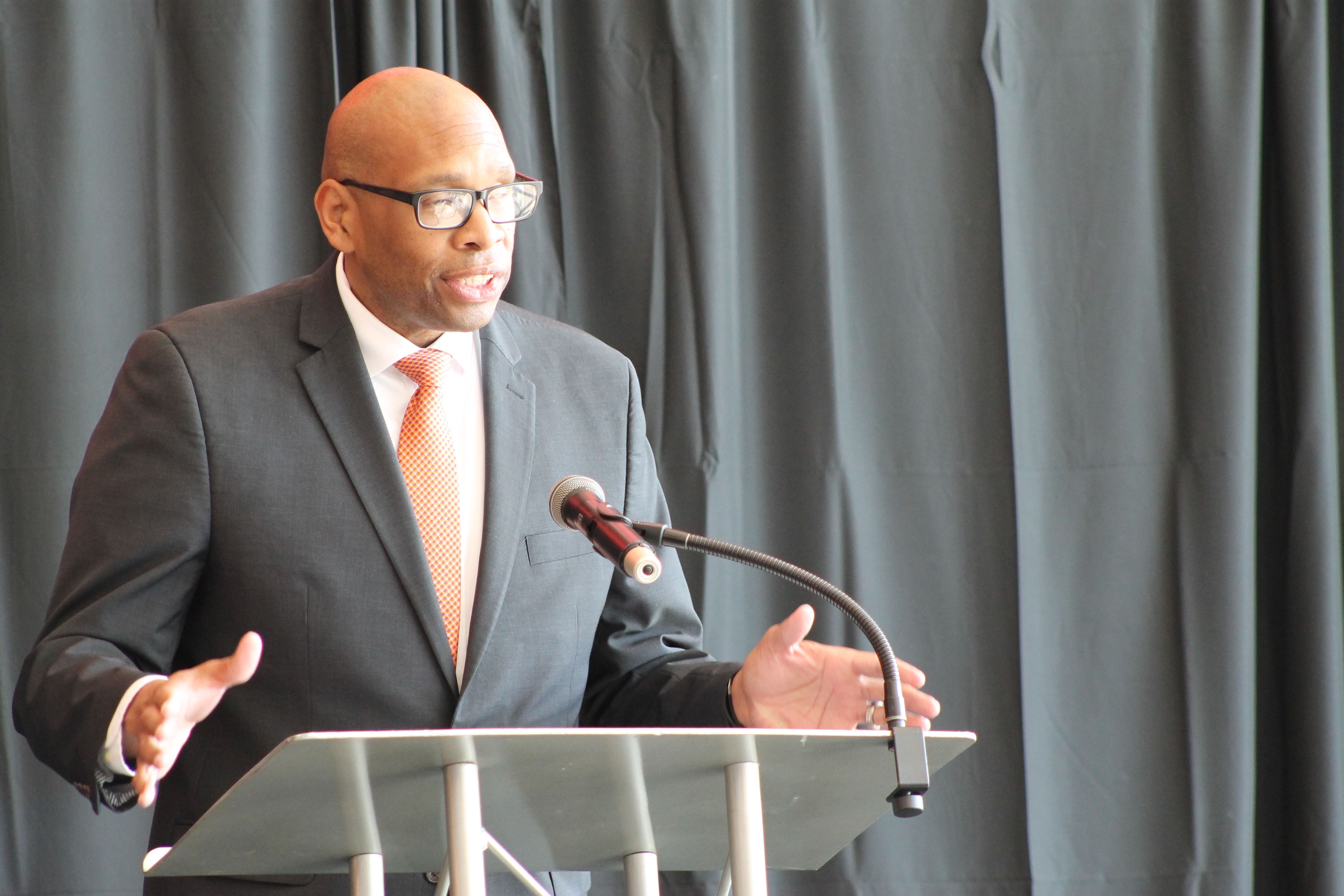 Bald man in dark suit, white shirt, and orange tie speaking at podium with microphone in front of dark curtain backdrop.