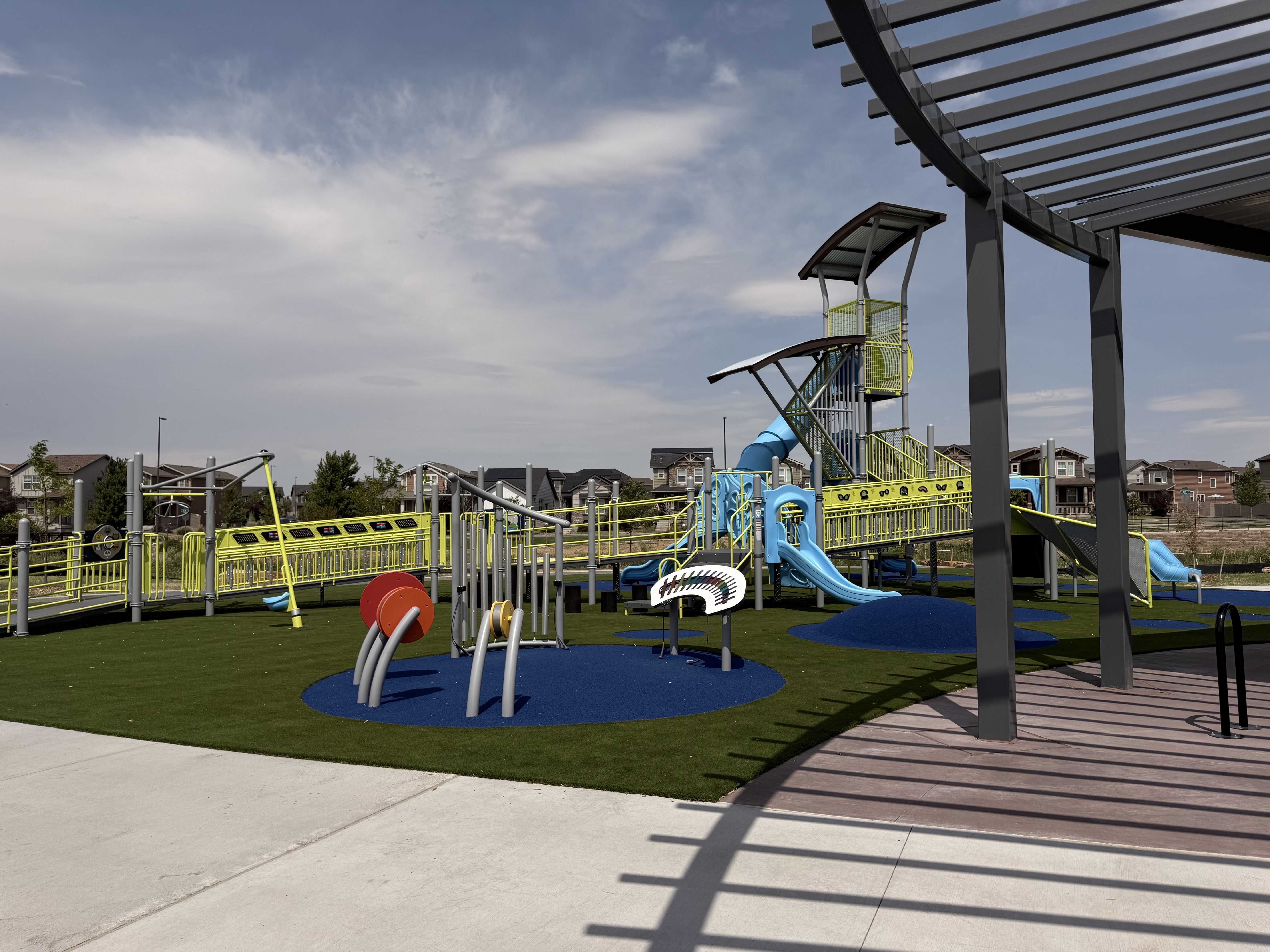 Colorful playground with blue slides and yellow rails, climbing structures on green turf. A gray pergola extends on the right; houses and a blue sky with clouds in the background.