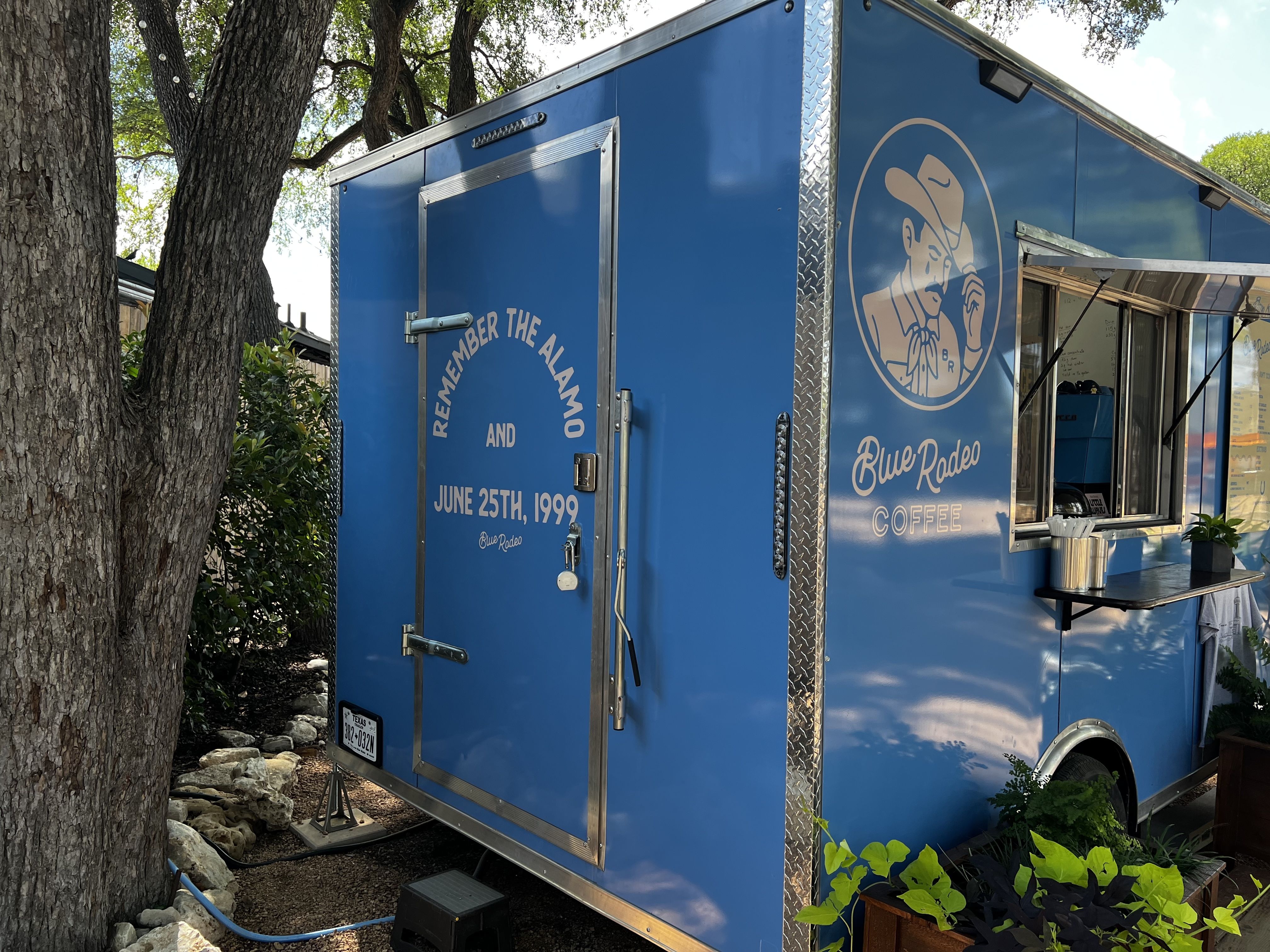 Blue food trailer with chrome trim and a large circular logo of a man in a hat, serving window open; outdoor setting among trees with a coffee stand.