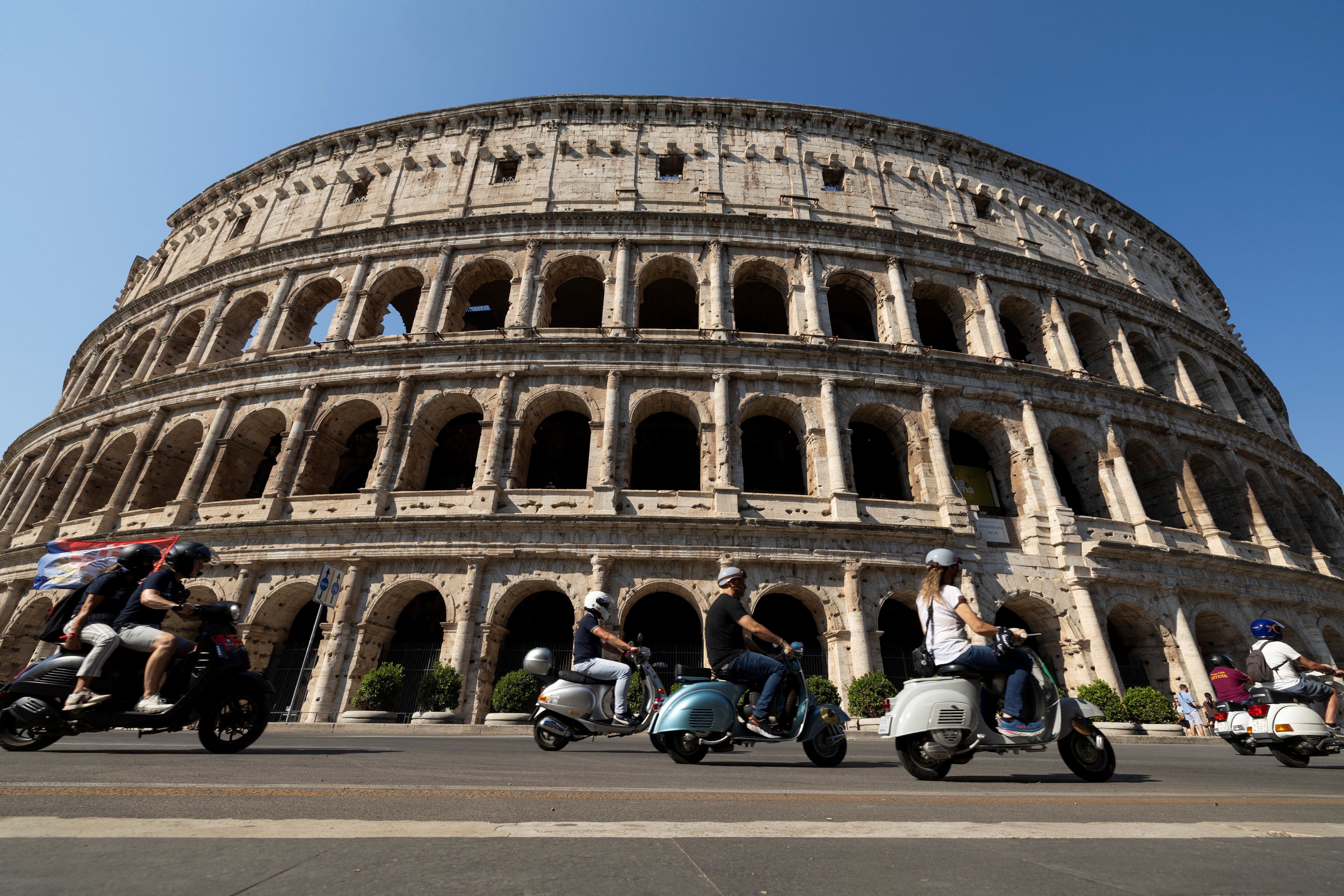 Side view of the ancient Colosseum with its stacked arches under a clear blue sky; a line of motor scooters and riders cruise in the foreground.