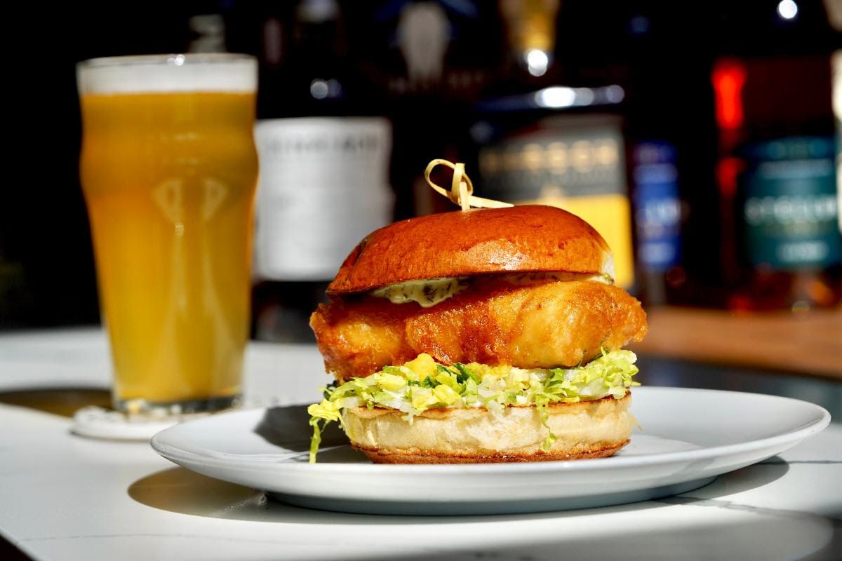 A crispy fish sandwich with a beer in the back