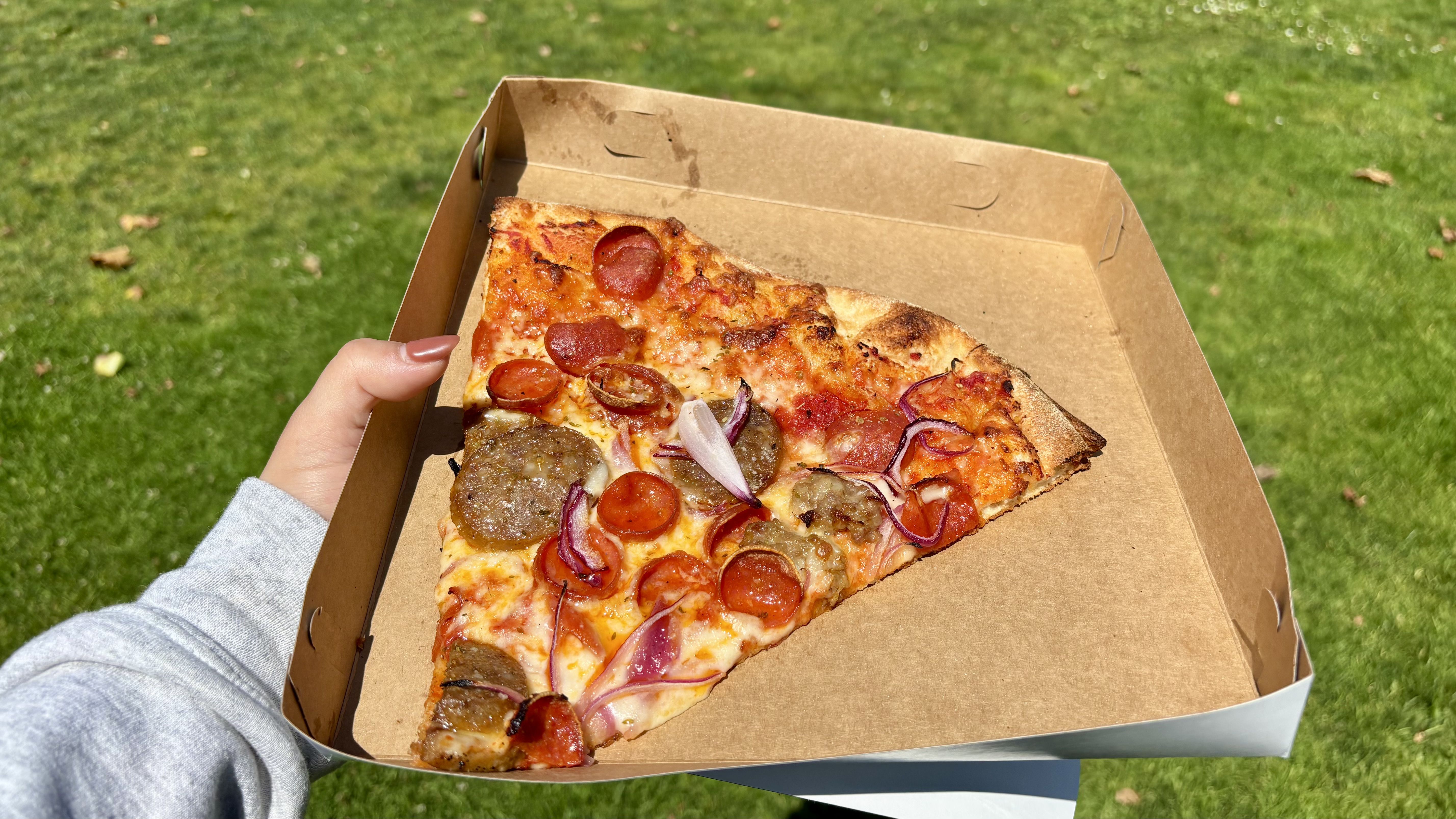 Hand holding a brown pizza box with a single slice of pizza topped with pepperoni, sausage, and red onions, against a green grassy background in sunlight.