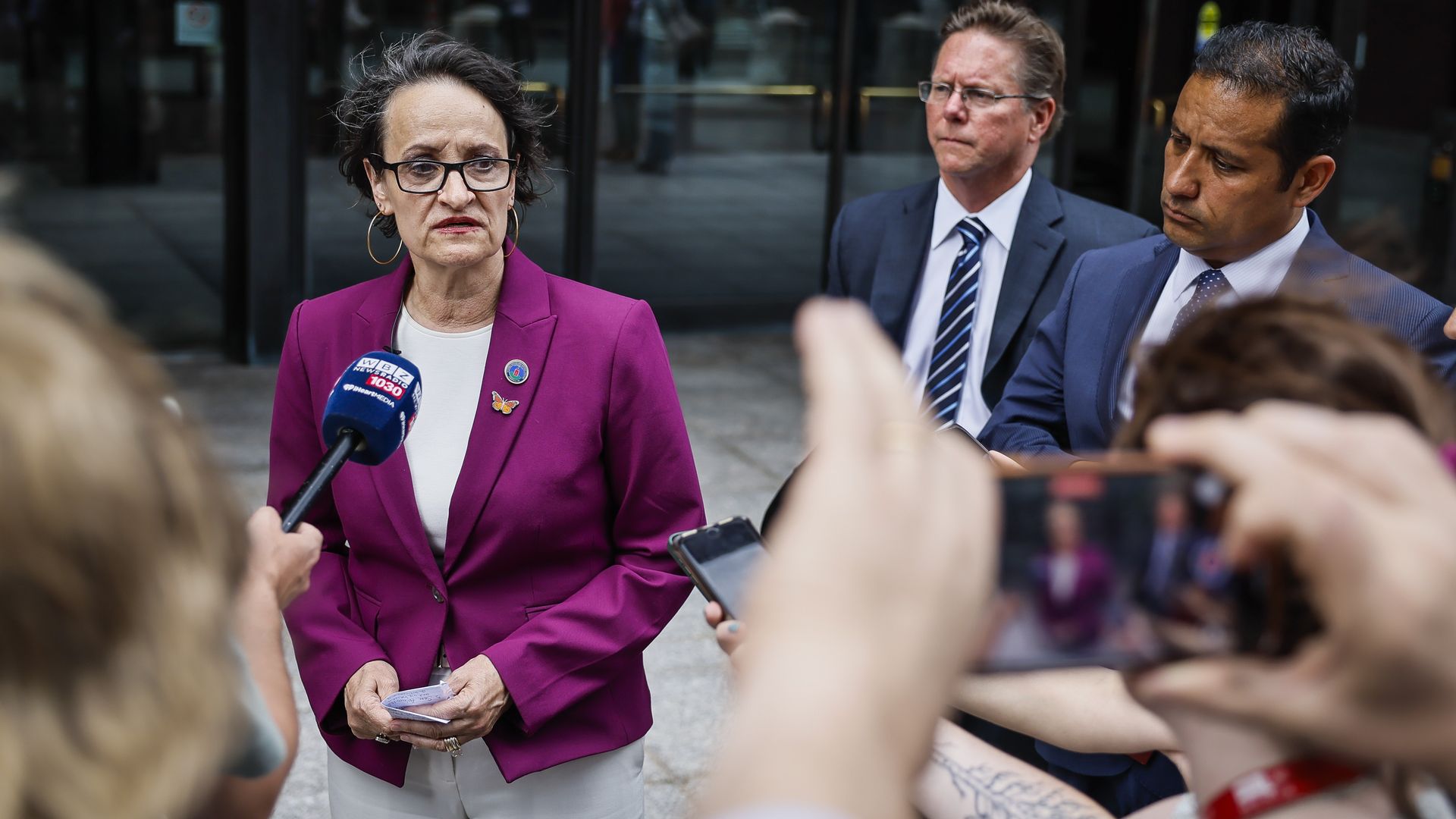 Woman in magenta blazer speaking to reporters holding a microphone and phone, two men in suits stand behind her, outside a building with glass doors.