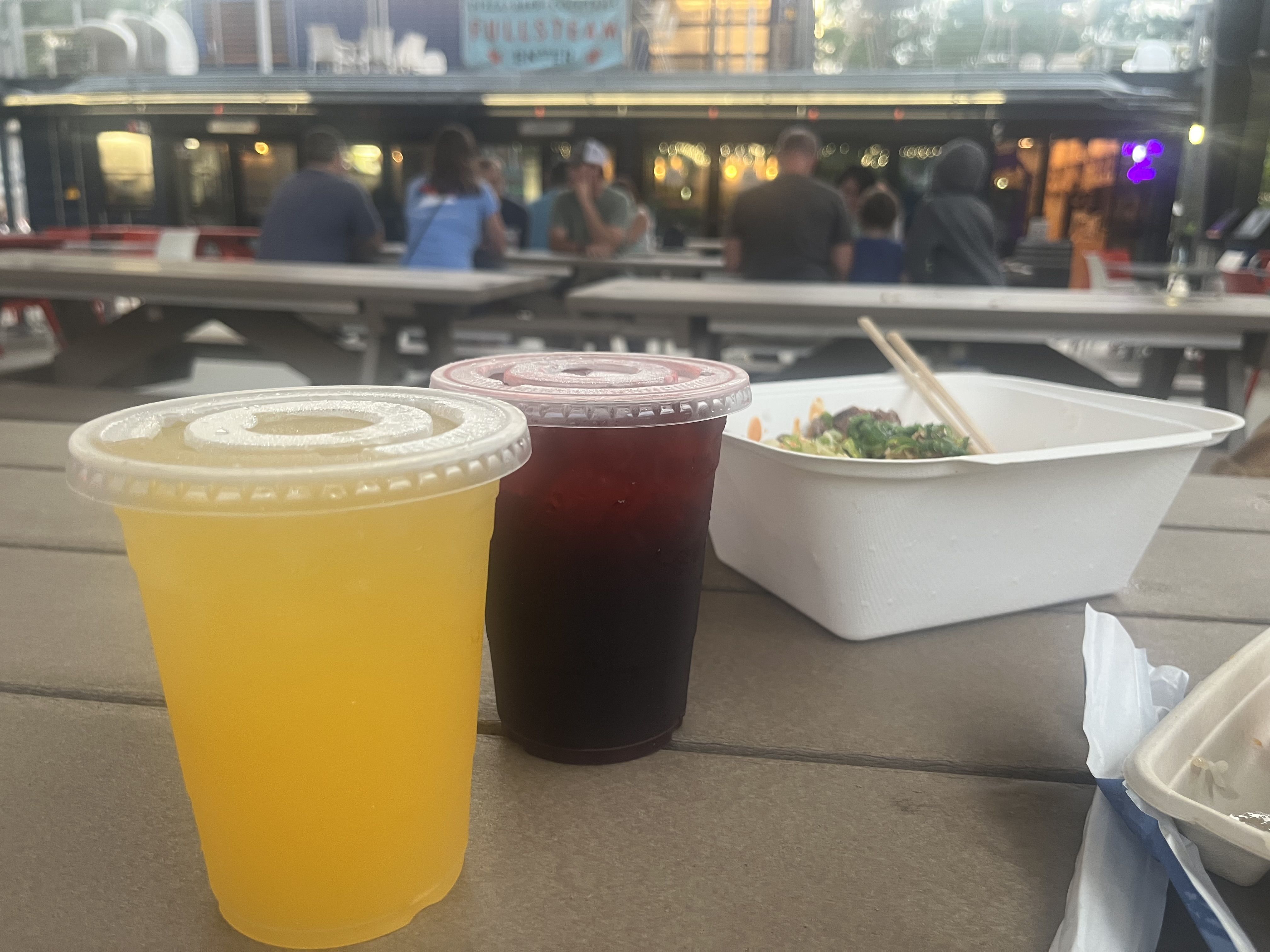 Two takeout cups with lids—yellow, purple—and a white container with chopsticks on a wooden table; blurred background shows people at an outdoor food hall near storefronts with warm lights.