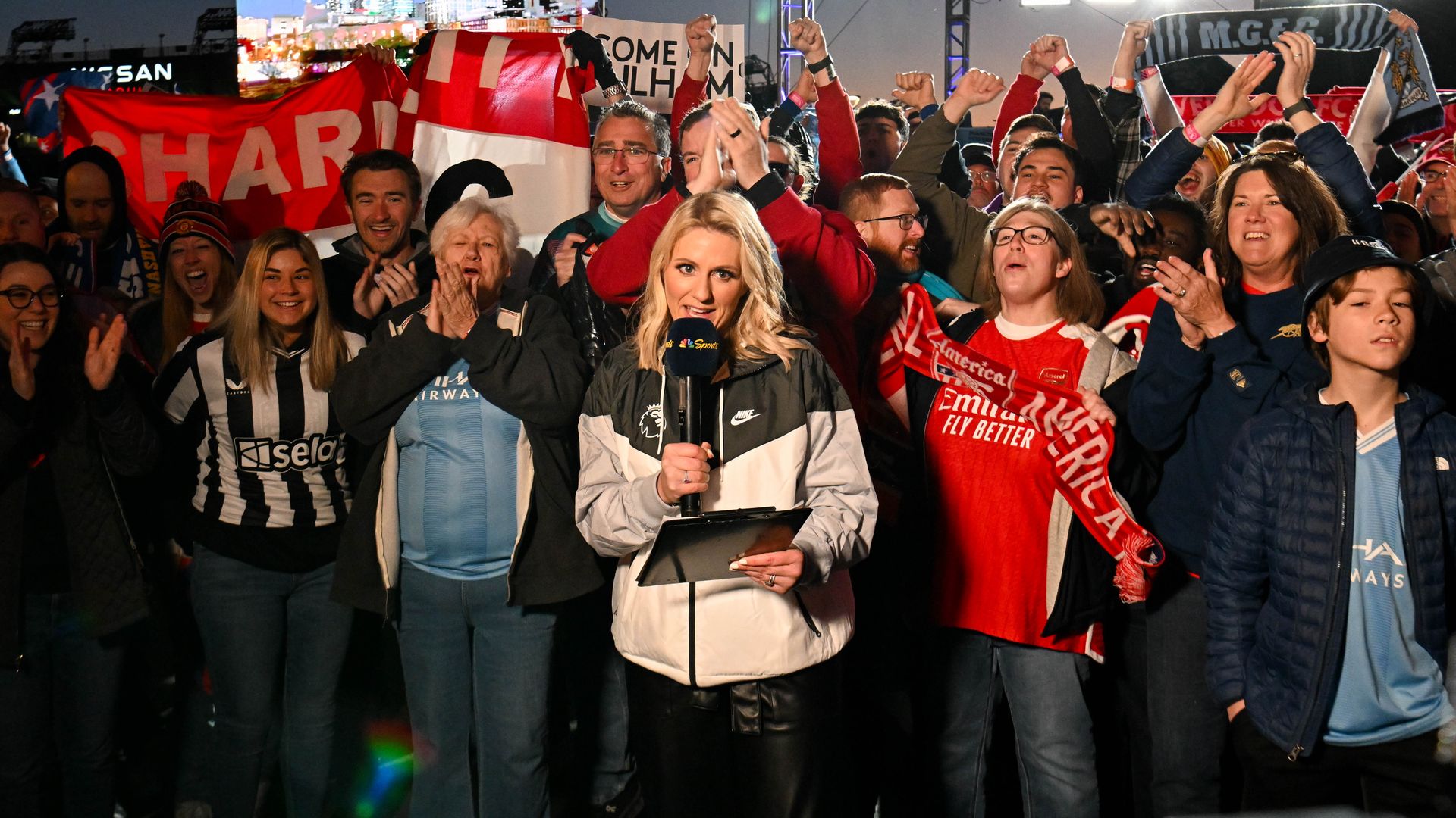 Large crowd at a stadium event, fans wave red-and-white flags as a female reporter with a microphone stands front and center; bright stage lights and a cityscape video screen glow behind them.