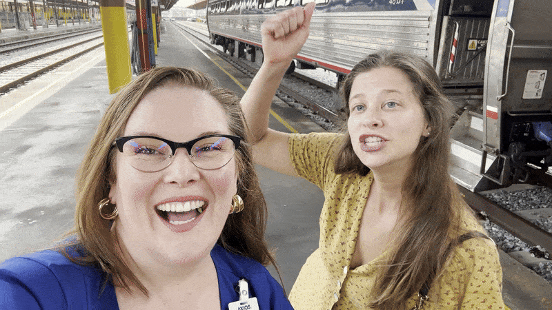 Two women at a train station platform, one in blue with glasses smiling at camera, the other in yellow with long hair raising her arm, a silver train is in the background.