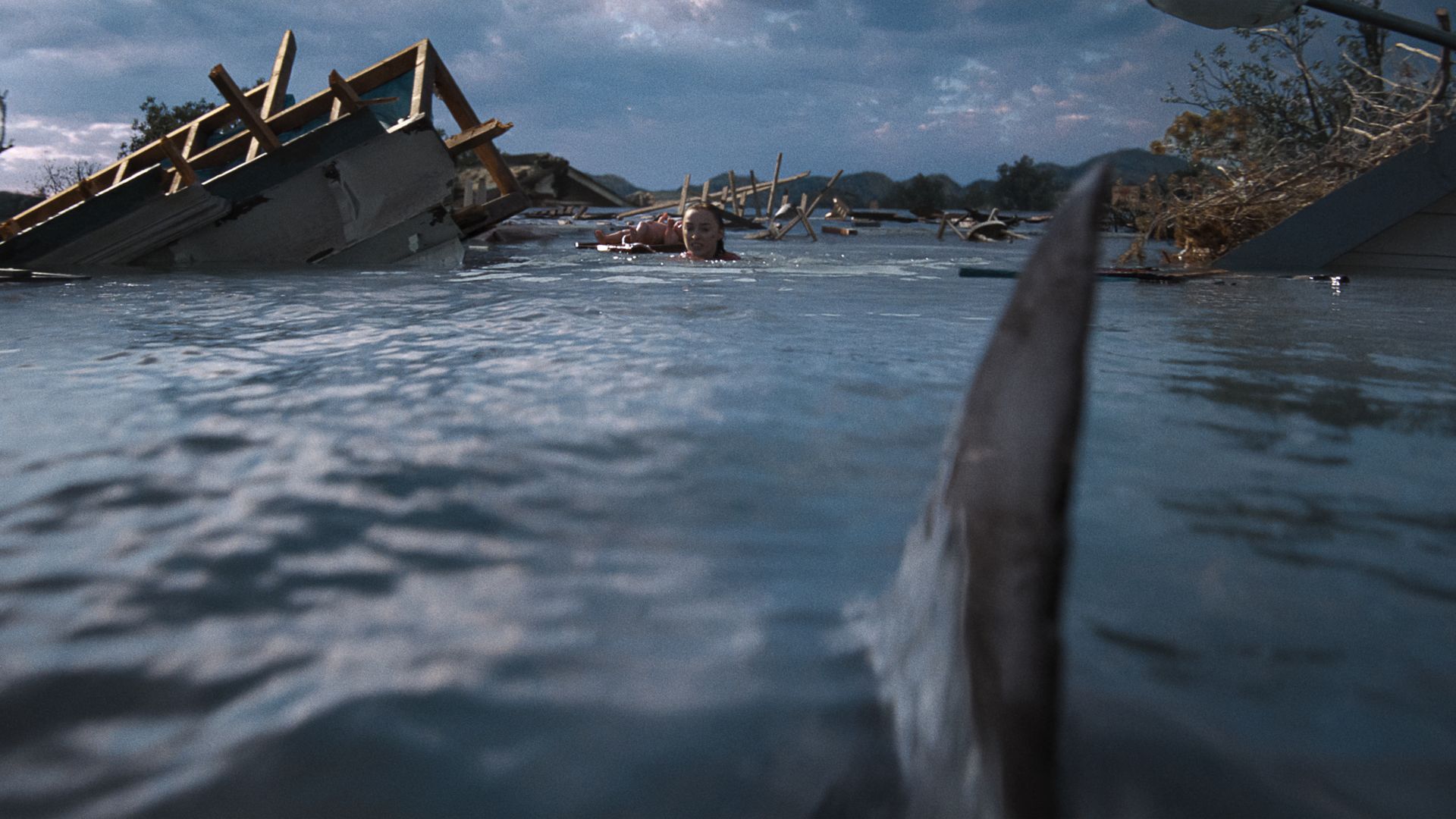 Rescue scene: a person with head above water swims among wrecked debris and submerged structures in a flooded neighborhood under a cloudy sky.