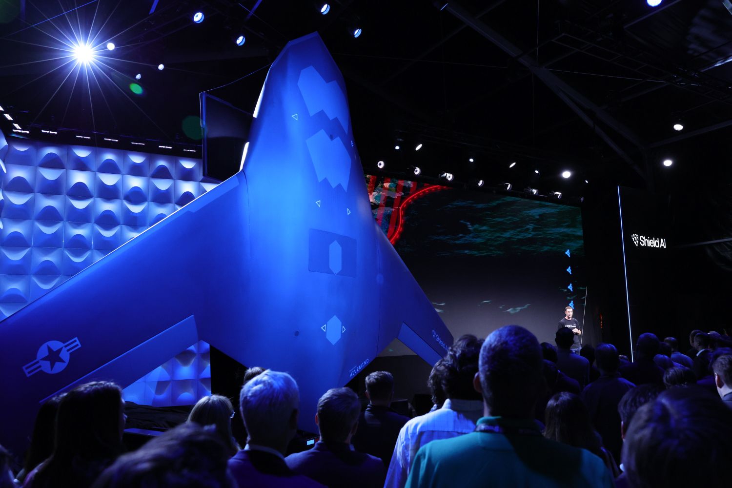 Large blue drone displayed at a tech event with an audience watching a speaker on stage. Shield AI logo visible on screen in a dark room lit with blue and white lights.