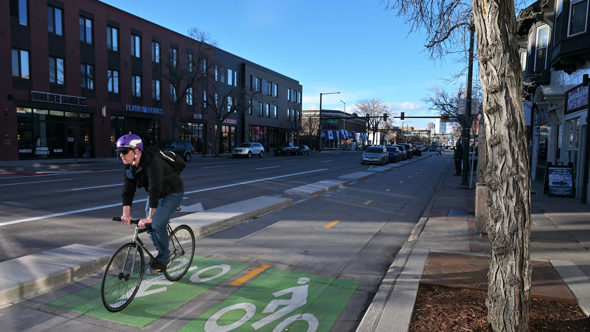 A person wearing a purple helmet rides a black bike on a green bike lane in a city street with shops and parked cars under a clear blue sky.
