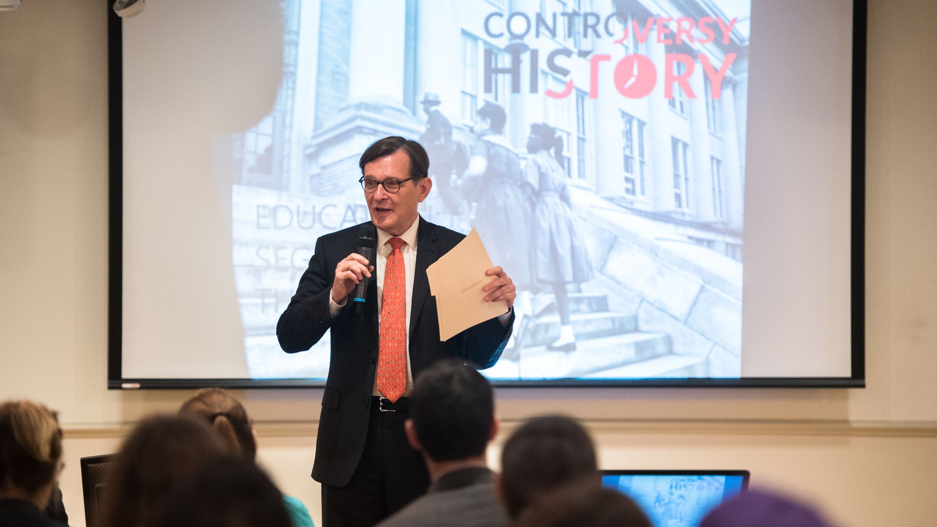 Man in a dark suit with an orange tie, wearing glasses, speaks into a microphone, holding papers. A projection screen behind shows a black-and-white city scene with red text "CONTROVERSY THIS STORY".