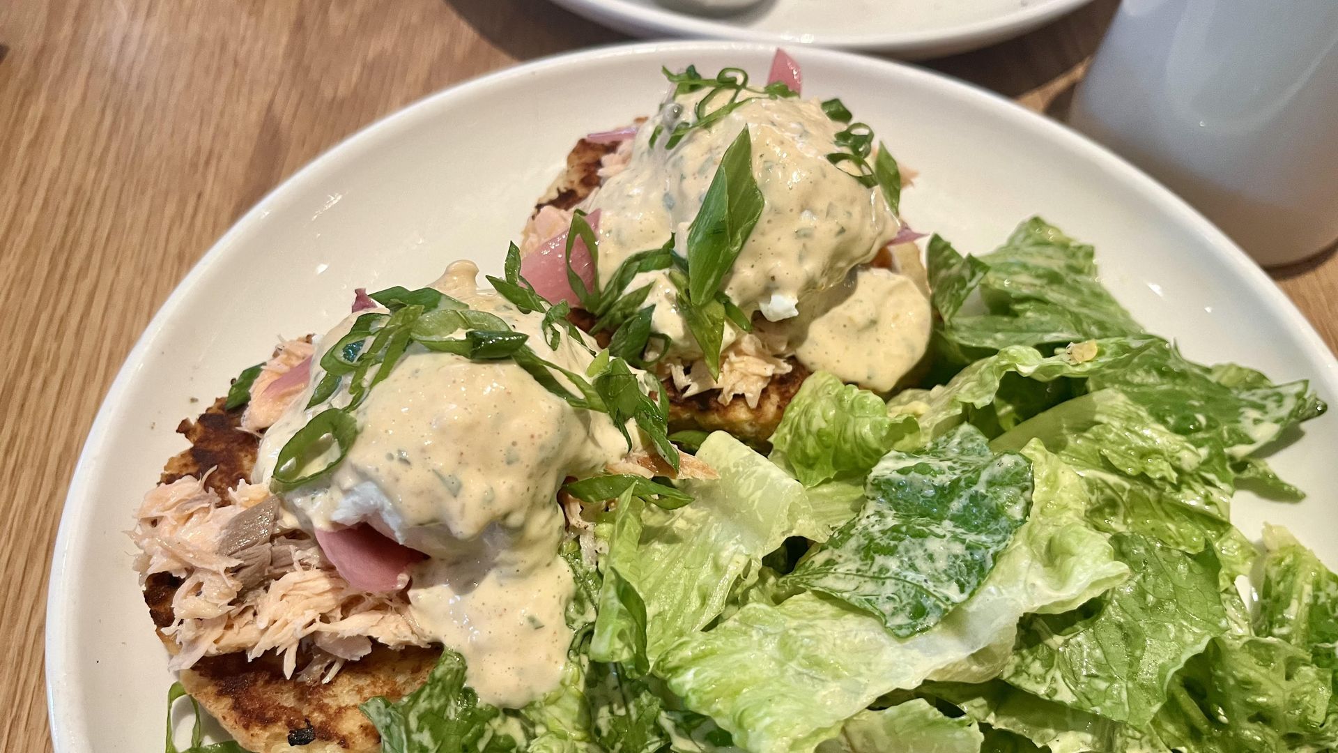 A round white plate with poached eggs and salmon and sauce on top of biscuits, with a green side salad.