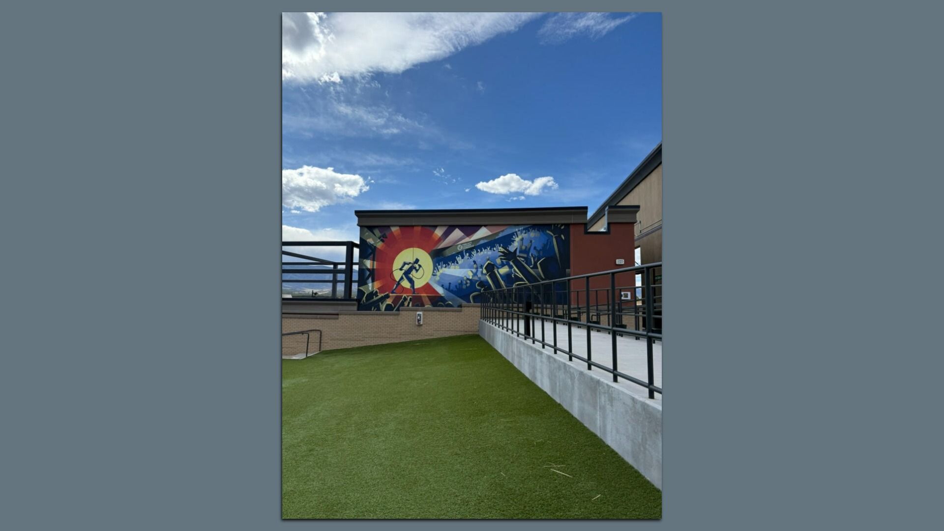 A mural in the same color scheme and shape of the Colorado state flag is painted onto a wall inside a space that features a green grass area and porch space. The sky is blue with with a few white clouds above. 