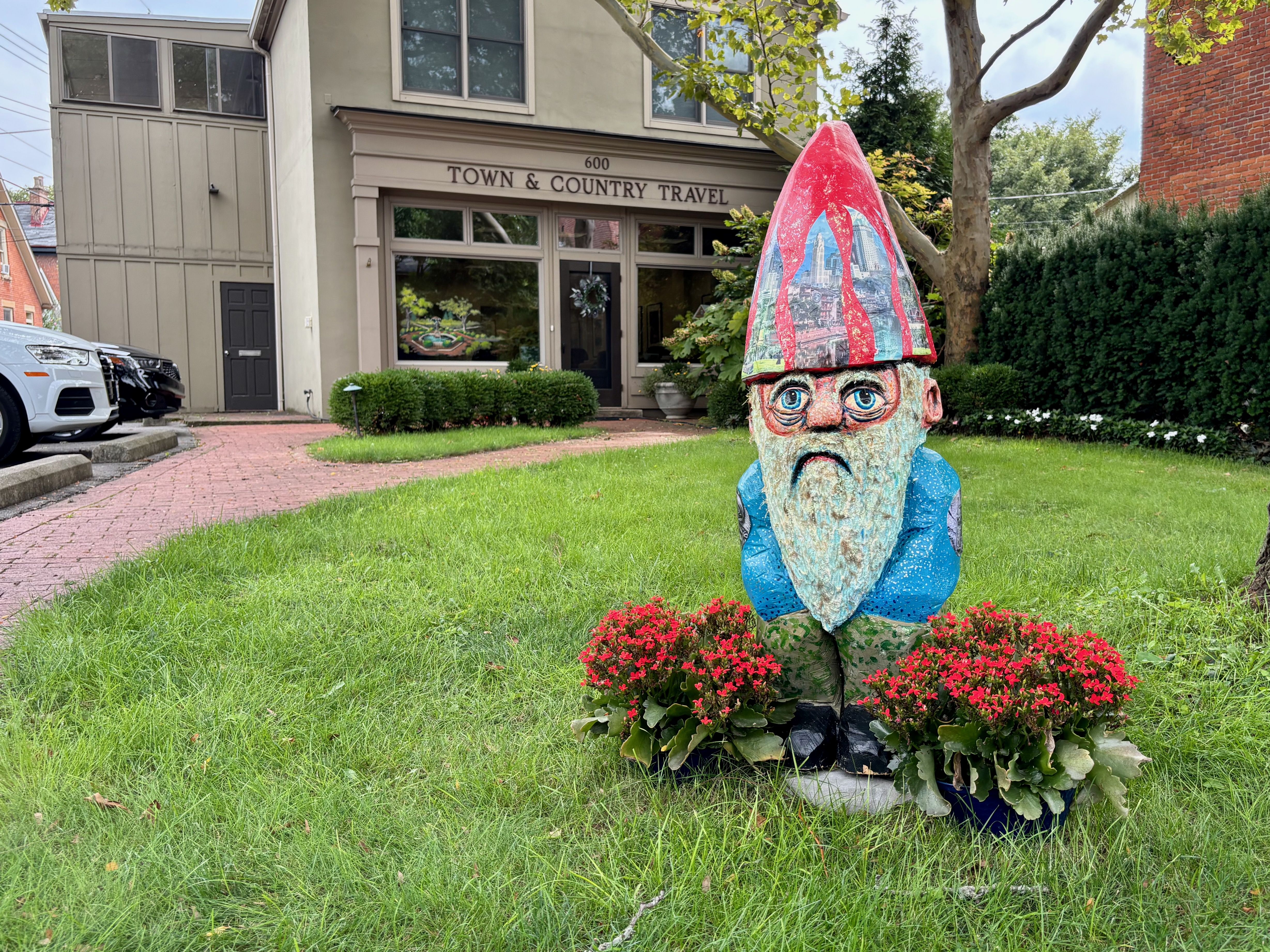 Storefront reading "Town & Country Travel" at 600 with a large painted garden gnome in a red hat featuring city skylines, on a green lawn beside trees and parked cars.