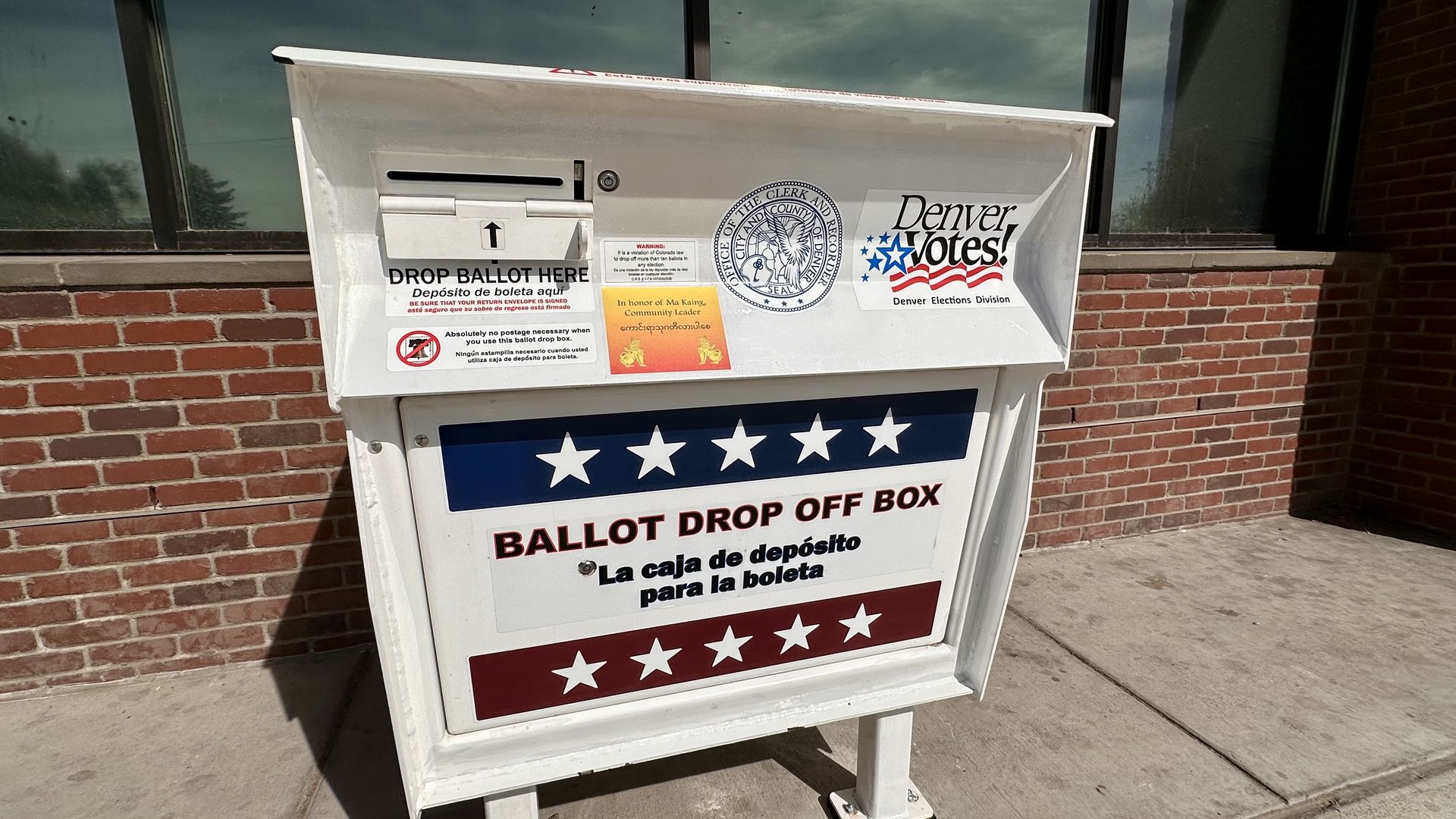 White ballot drop-off box outside a brick building; stickers read DROP BALLOT HERE and Denver Votes. A blue star stripe decorates the front with a round seal nearby.