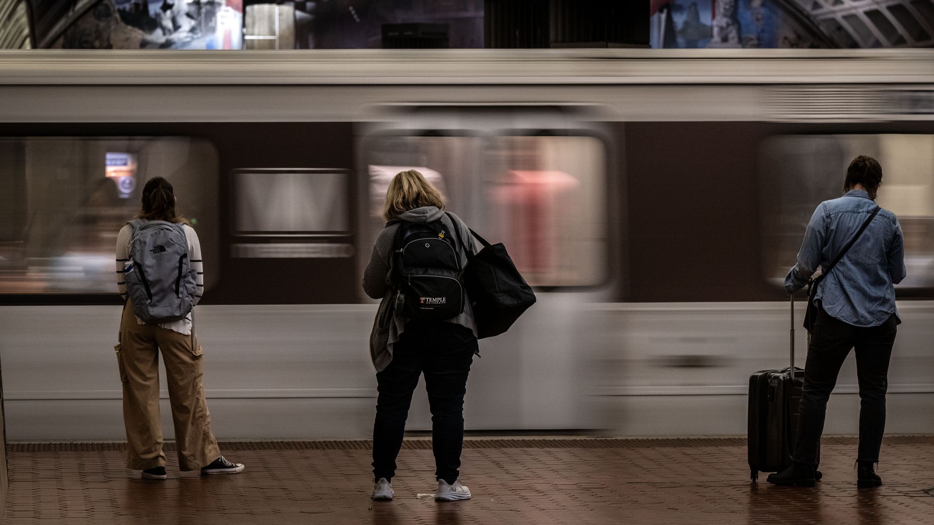 Three women stand six feet apart waiting for a Metro train as it zooms by.