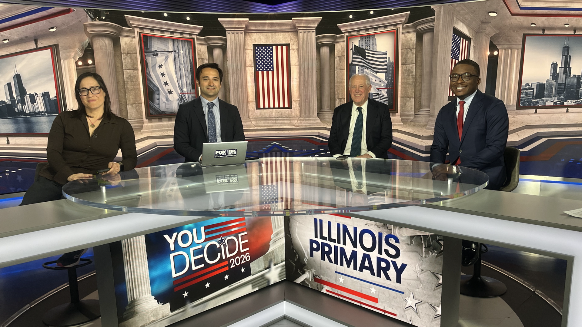 Four news hosts sit at a glass studio desk against a patriotic backdrop with columns, flags, and cityscapes. On screens read "YOU DECIDE 2026" and "ILLINOIS PRIMARY" in red, white, and blue.