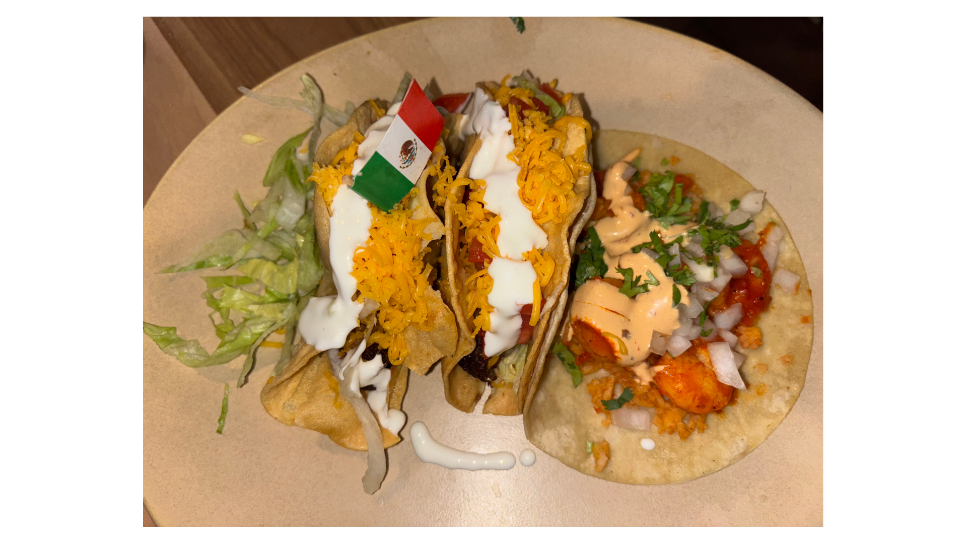 Plate of three tacos with meat, crema, shredded cheddar, lettuce, onions, cilantro and salsa; a small Mexican flag decorates the middle taco.