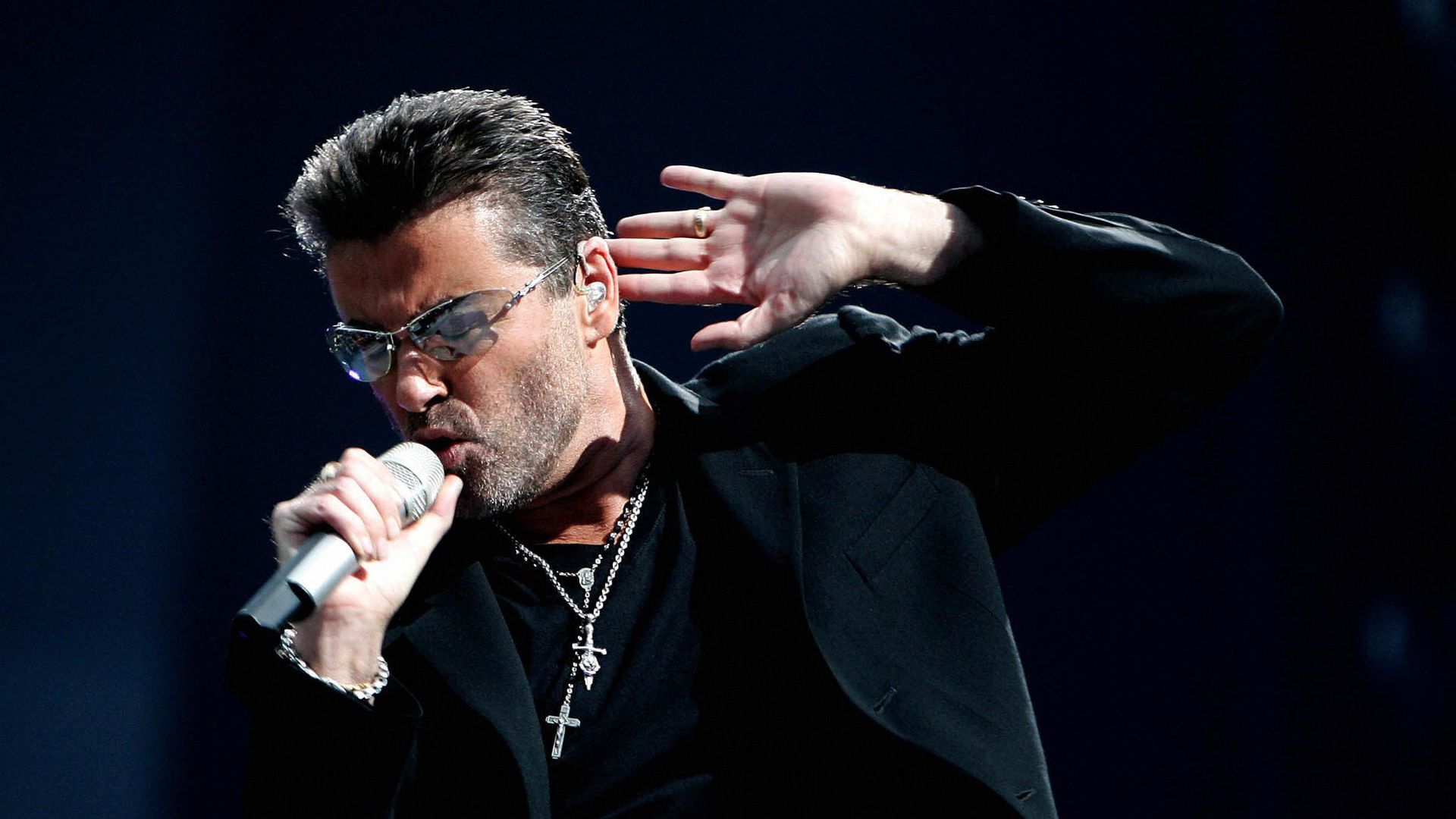 Late musician George Micheal sings into a mircrophone with his other hand near his ear. 
