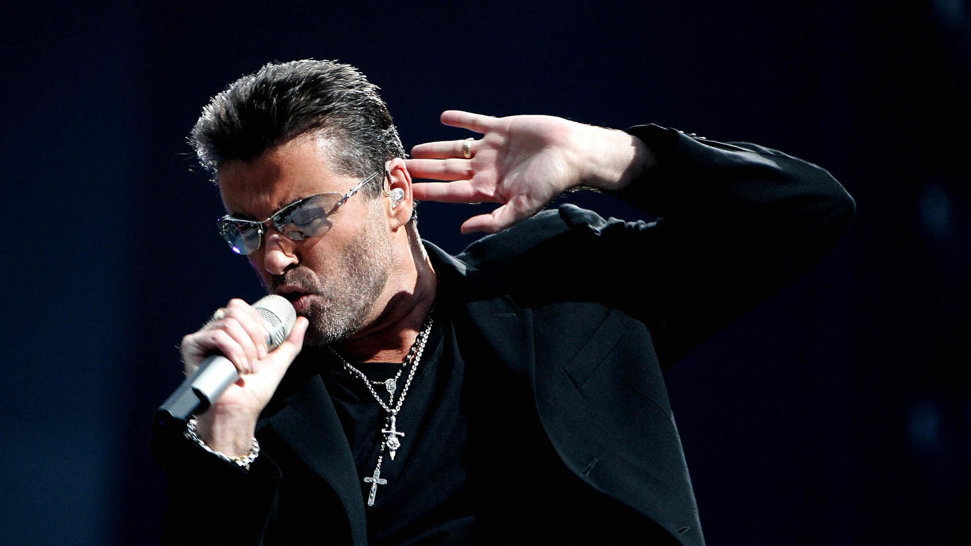 Late musician George Micheal sings into a mircrophone with his other hand near his ear.