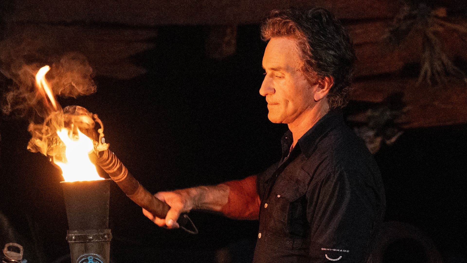 A man with a black shirt lighting a fire torch in a dark setting, with flames and smoke visible.
