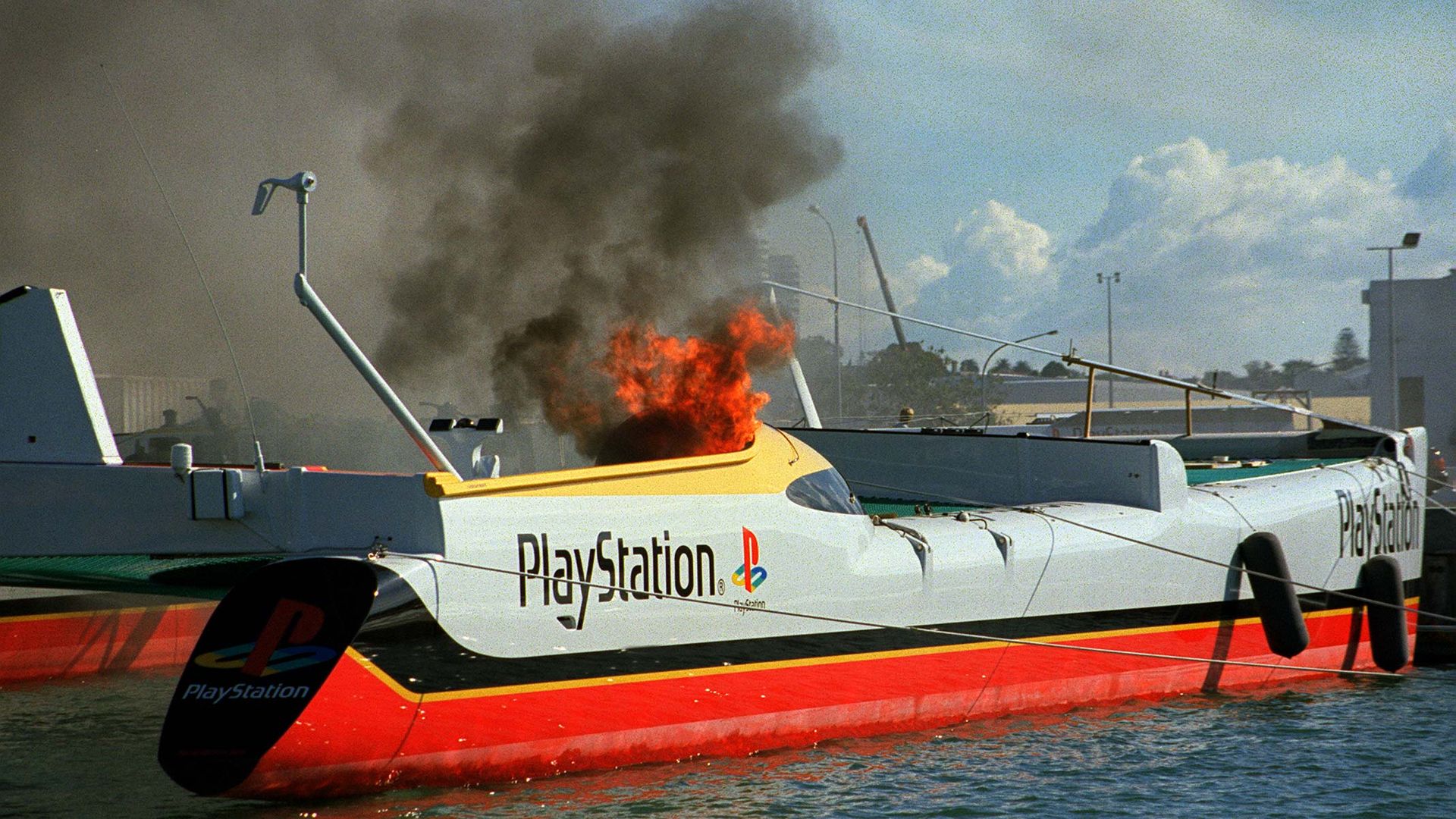 Photo of a catamaran on fire while docked in a harbor. The side of the vessel bears the PlayStation logo