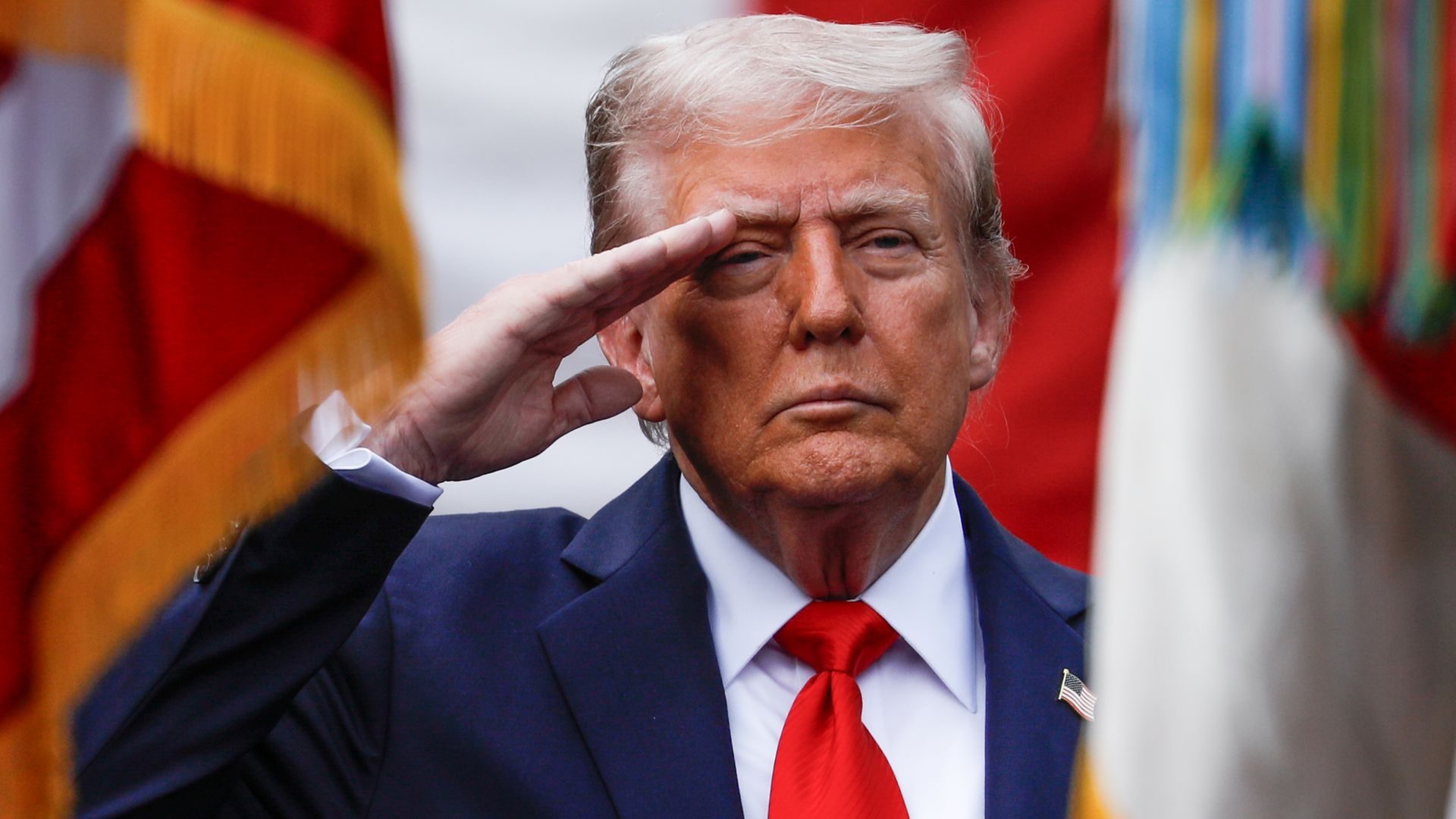 President Trump, in a blue suit and red tie, salutes. Behind him is a row of flags.