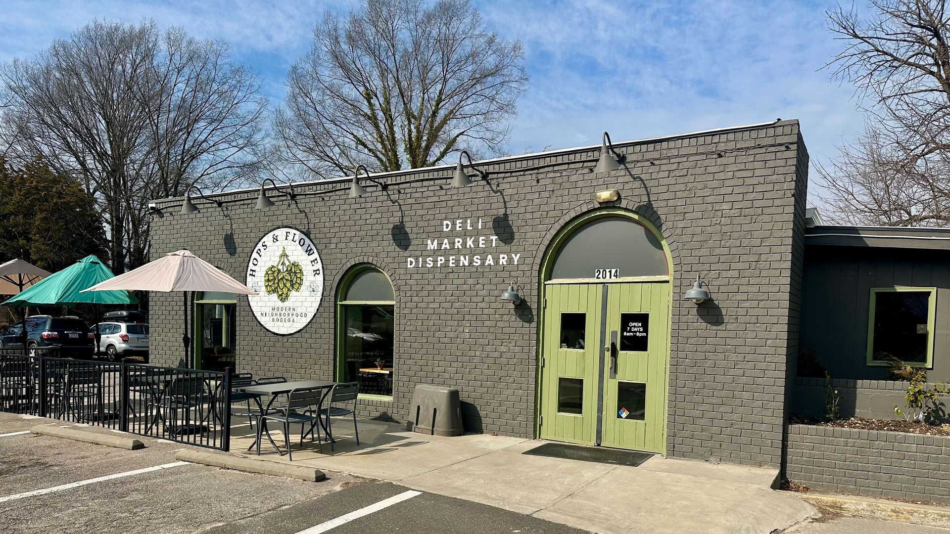 Gray brick building with green double doors labeled 2014, sign reads "Hops & Flower Modern Neighborhood Bodega," outdoor tables with umbrellas, and parking spaces in front.